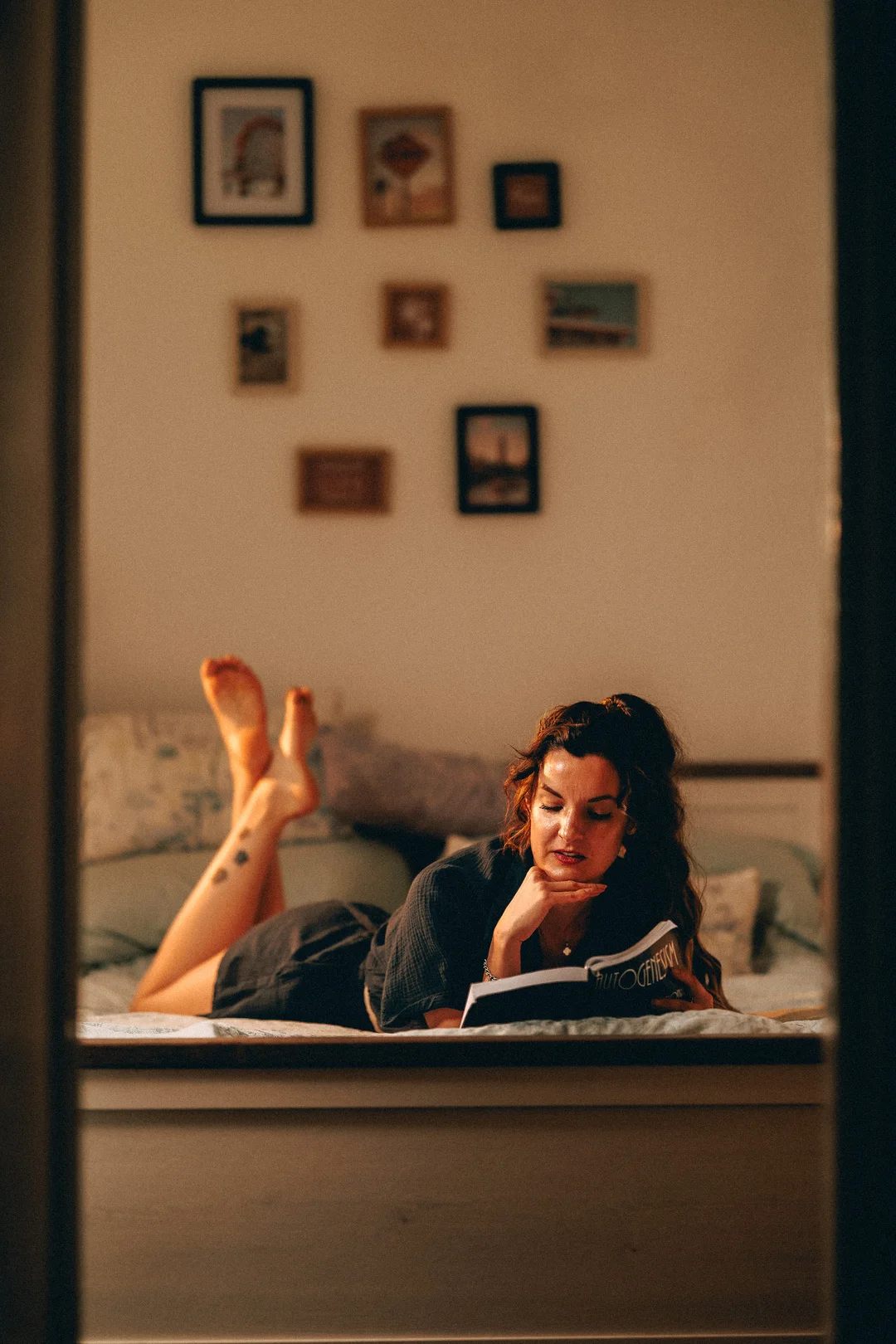 A woman lying on a bed reading a book in a cozy, dimly lit room with framed art on the wall.