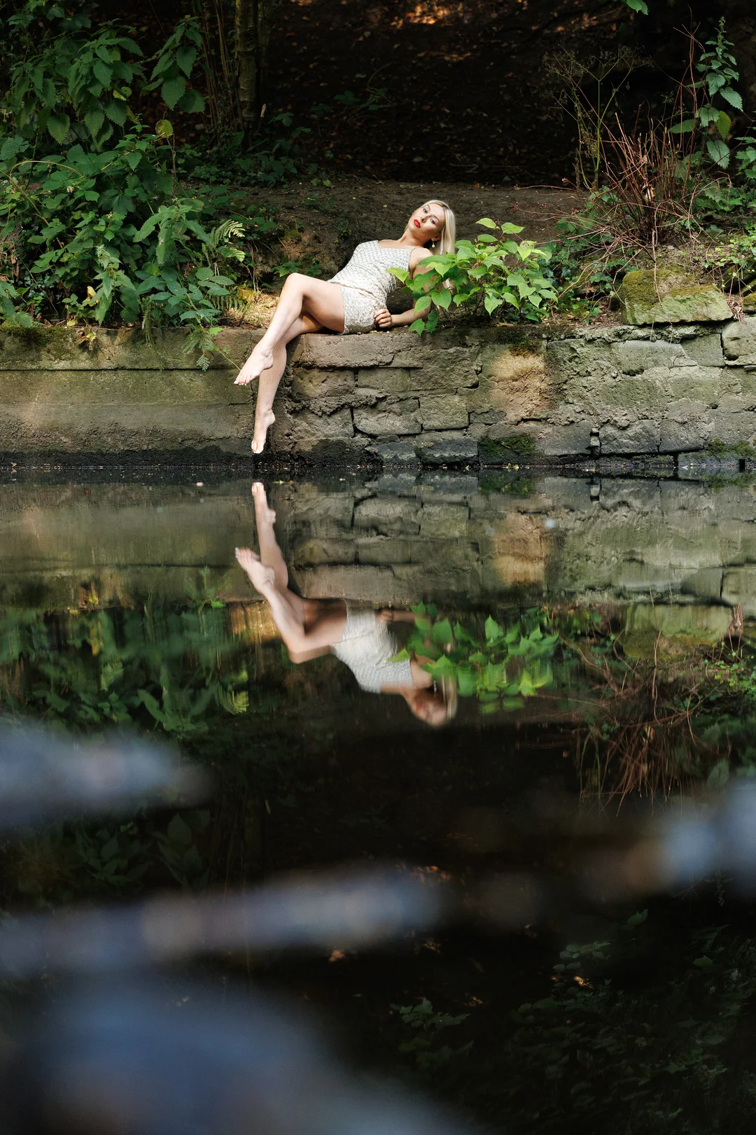 Woman in a gold sequin dress reclining on a stone wall by a pond, reflected in the water.