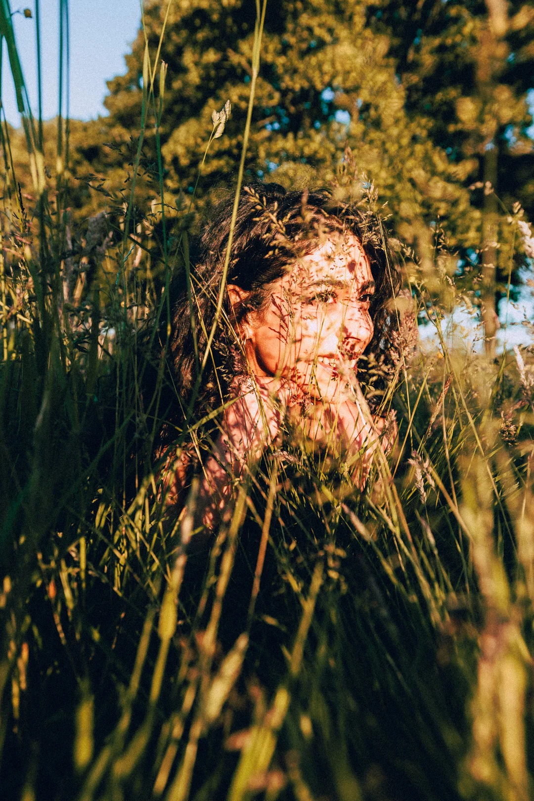 A woman's face partially hidden by tall golden grass in a sunny outdoor field.