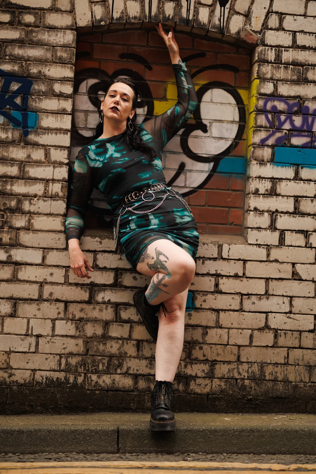 A person with tattoos in a teal and black dress posing against a graffiti brick wall.
