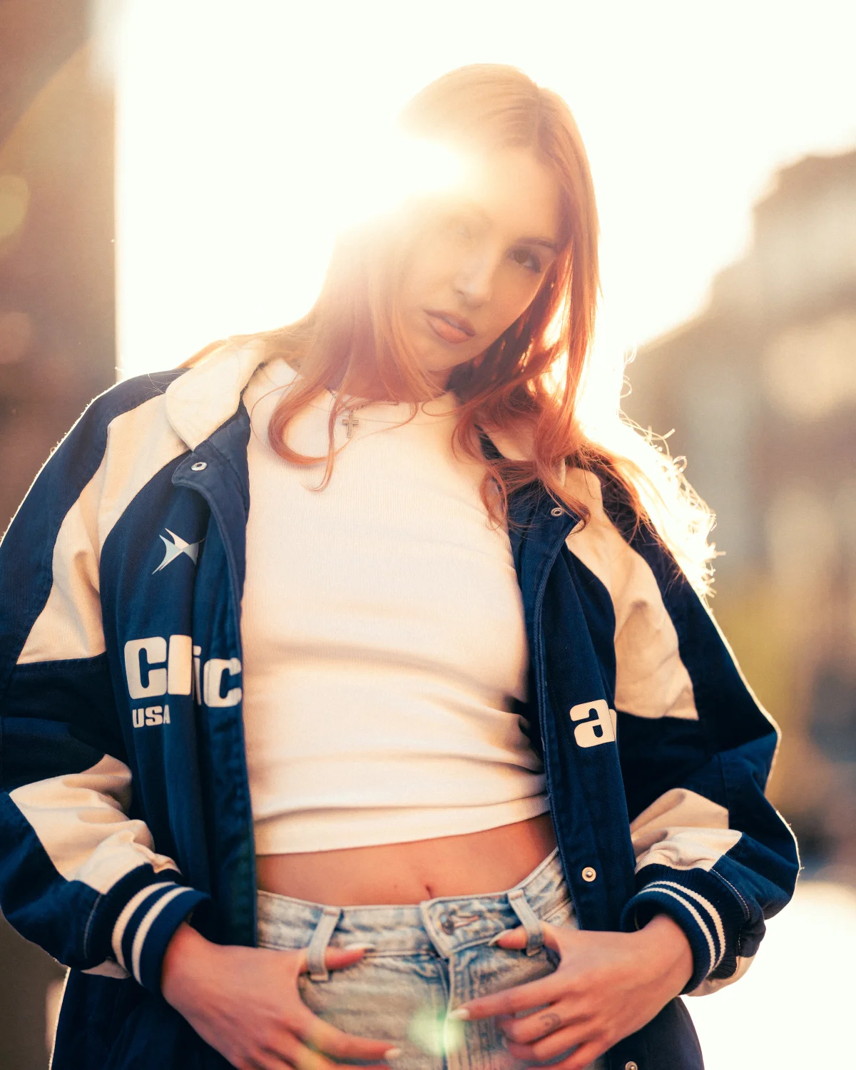 Young woman with red hair posing in a blue and white oversized jacket and white crop top, backlit by bright sun.