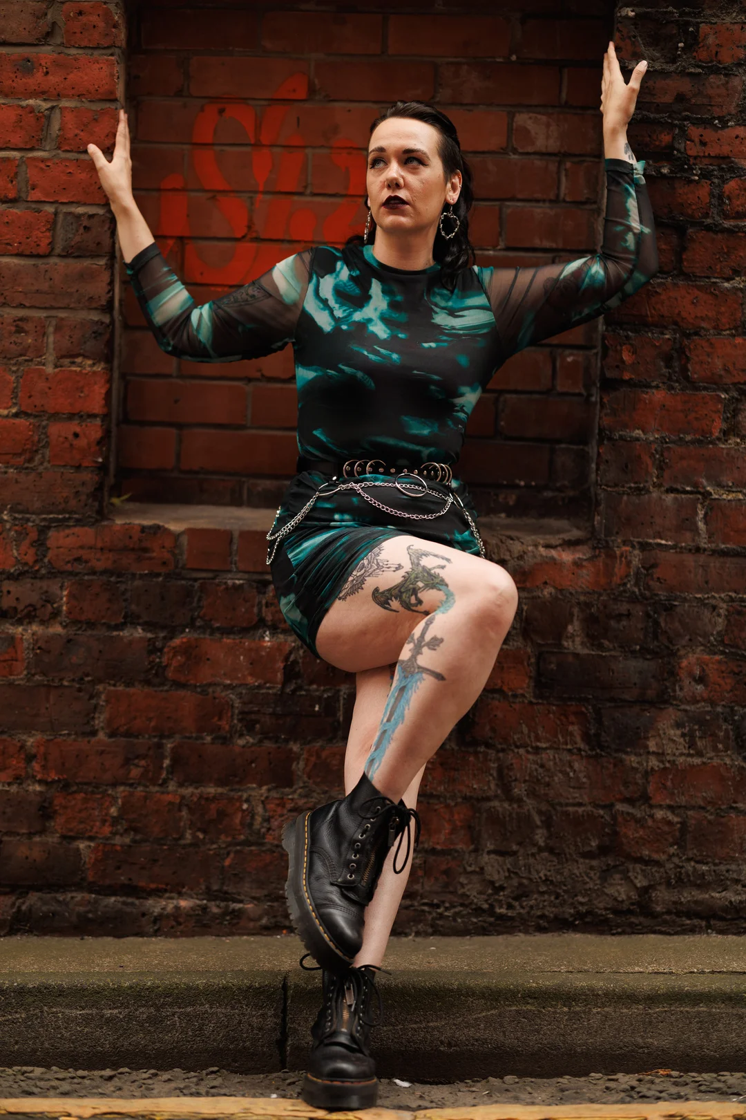 A woman in a teal and black patterned mini-dress and black boots posing against a red brick wall.