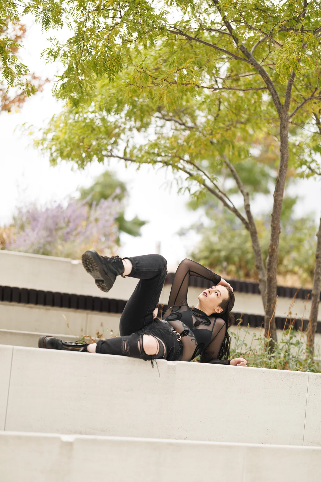 Woman in a sheer black top and ripped jeans posing on concrete steps under a leafy green tree.