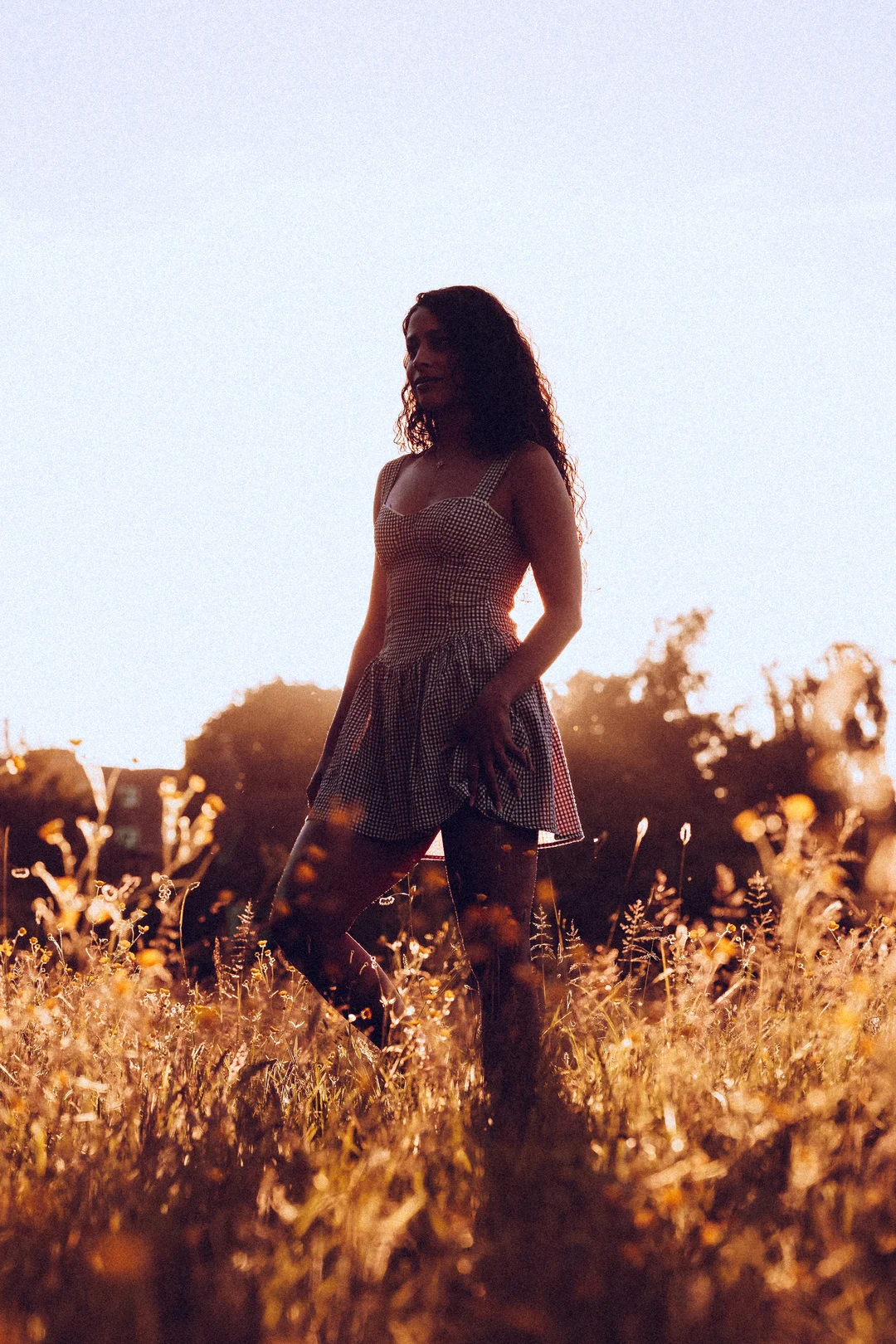 Woman with curly hair in a checkered dress standing in a golden field during sunset.
