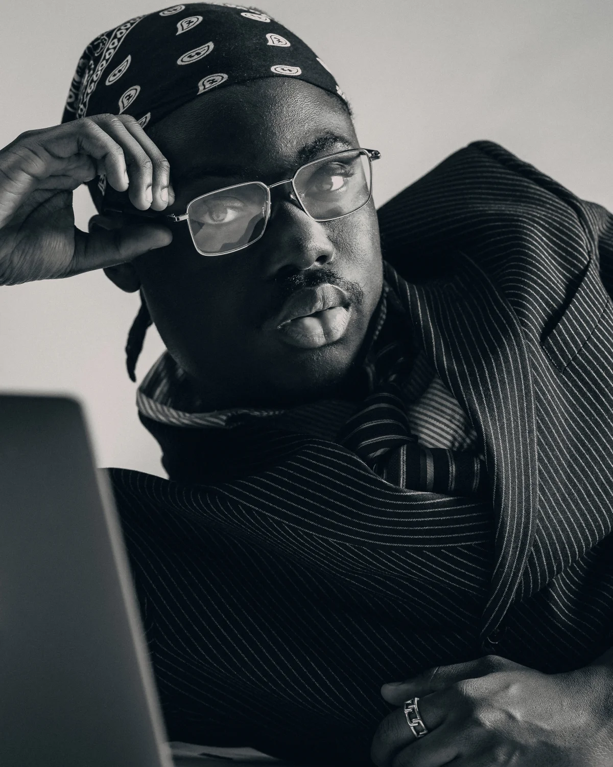 Black and white close-up portrait of a man in a pinstripe suit, glasses, and a bandana with a moody expression.