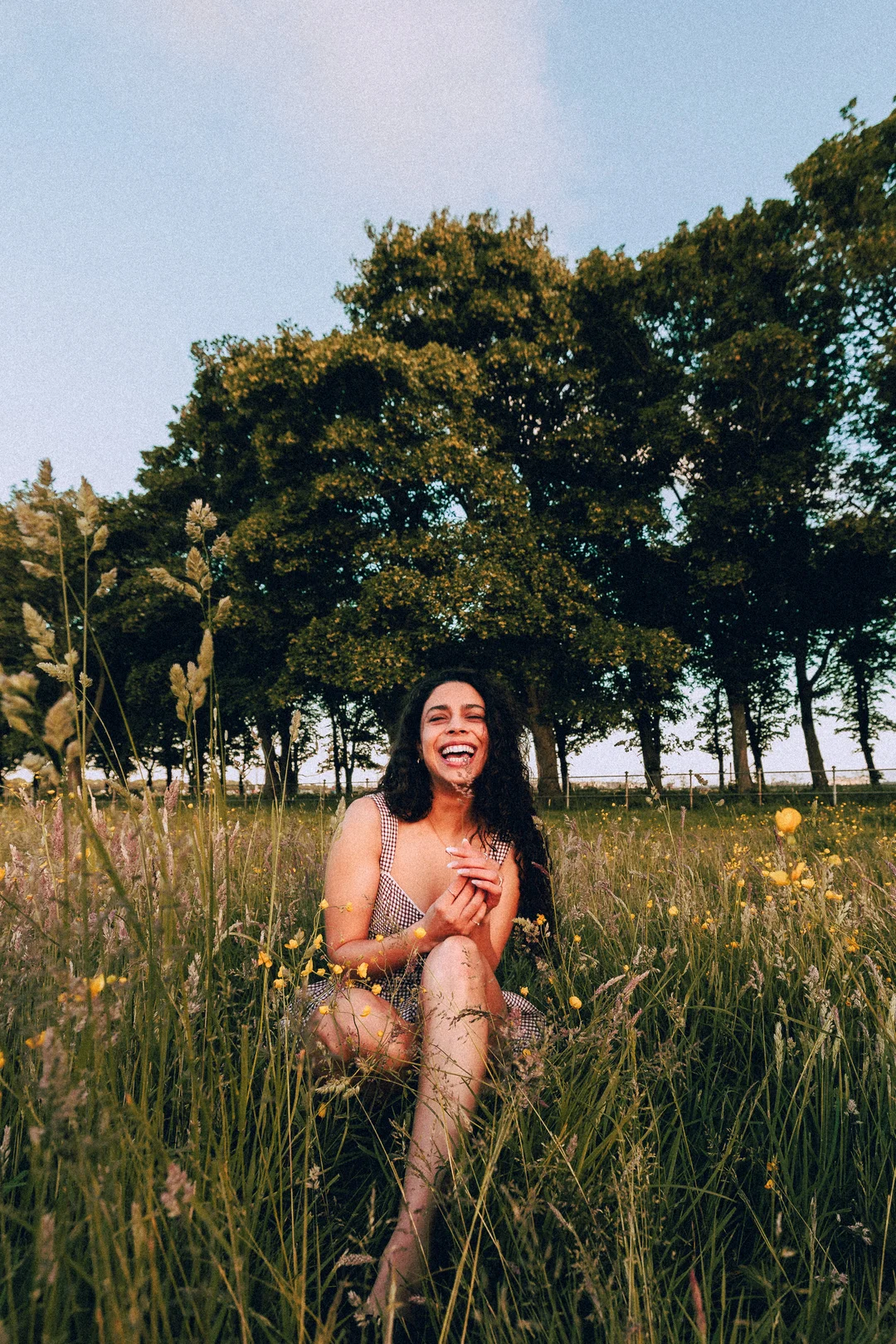 A joyful woman with curly hair laughing while sitting in a sunny grassy field with trees in the background.
