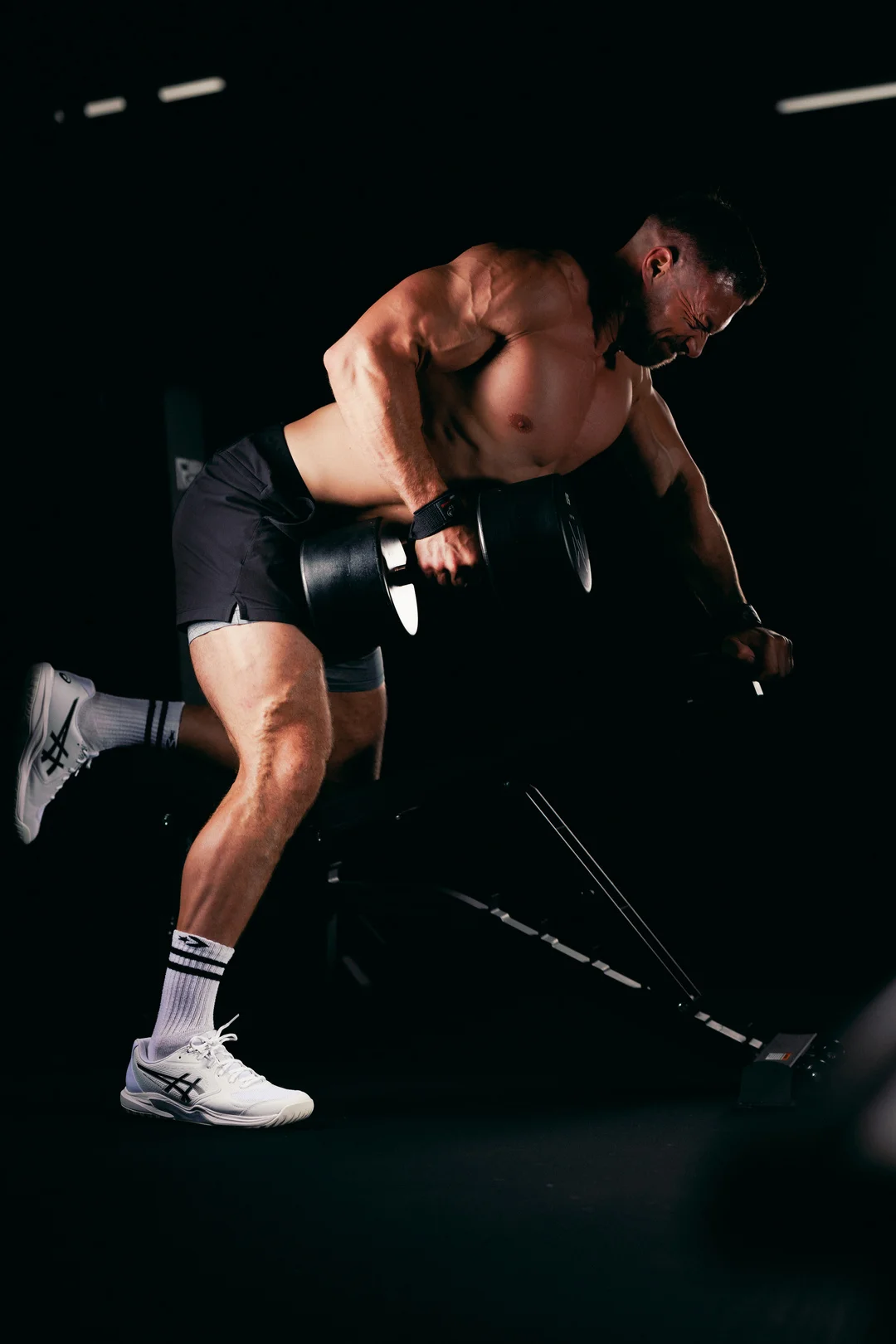 Muscular man lifting a heavy dumbbell in a dark gym, capturing intense physical effort.