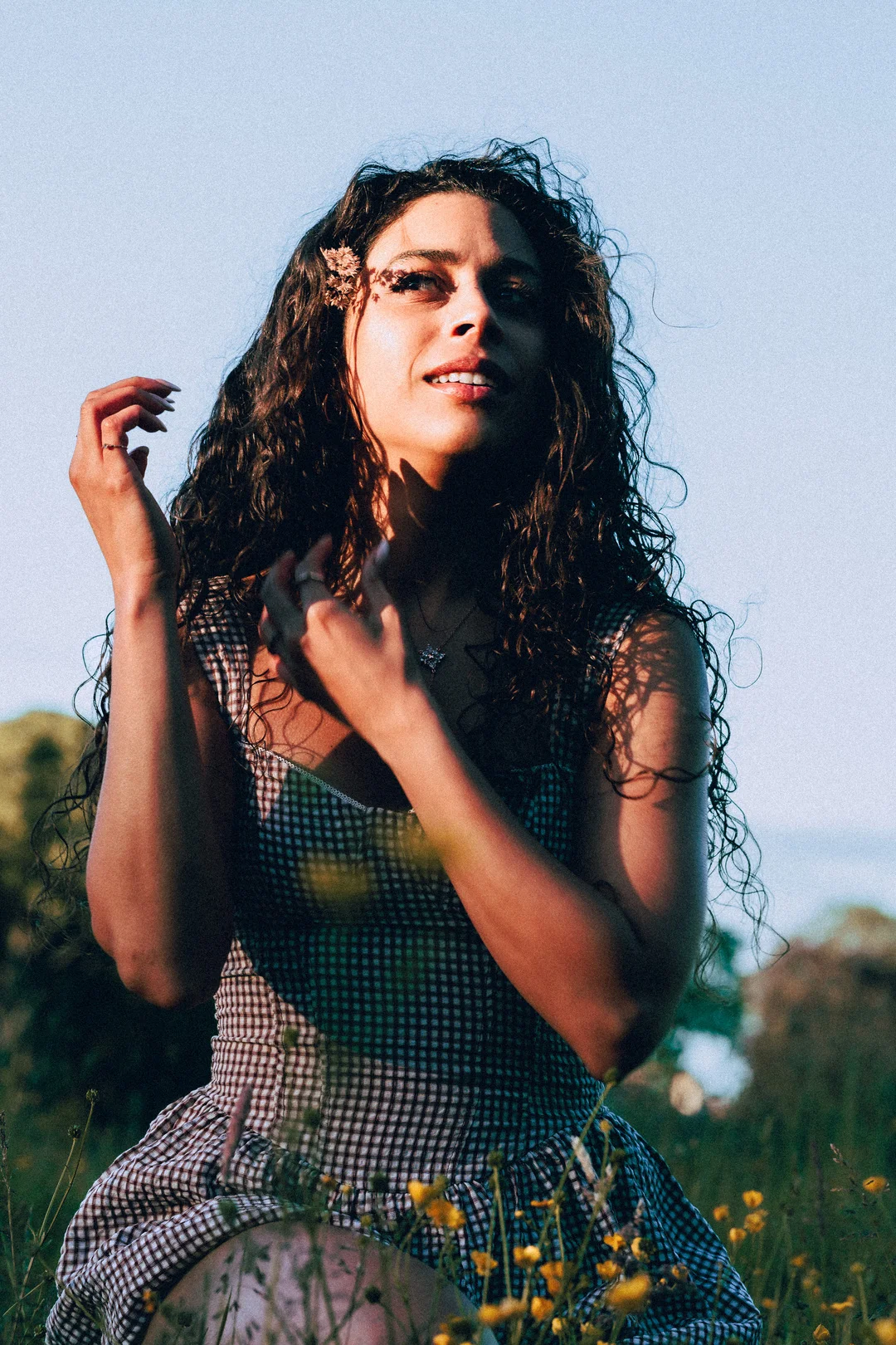 Woman with curly hair in a gingham dress sitting in a field of yellow flowers during golden hour.