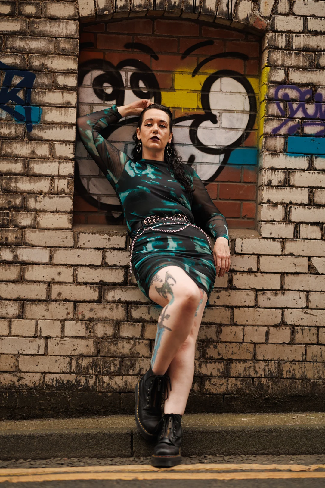 Woman with tattoos posing in a teal and black dress against a graffiti brick wall.