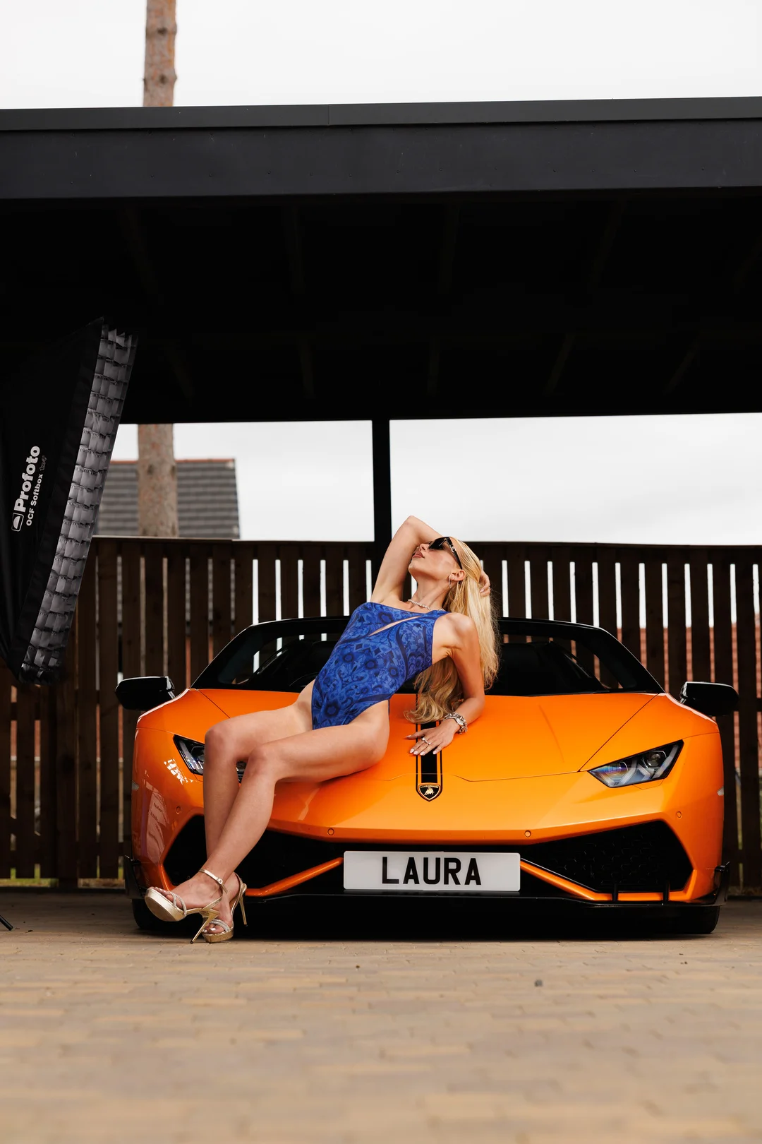 Blonde woman in a blue swimsuit posing on the hood of an orange luxury sports car.