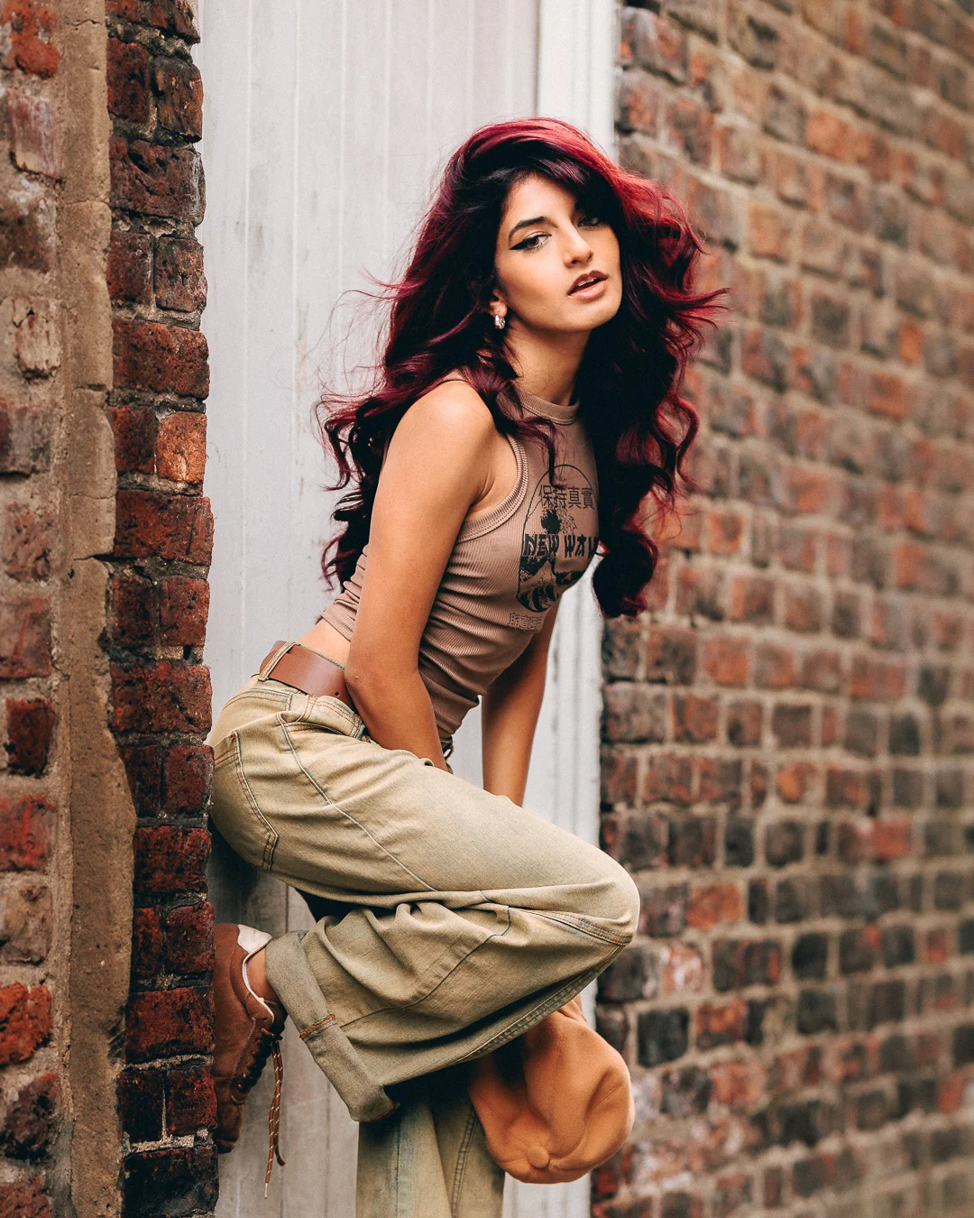 A woman with vibrant red hair posing against a brick wall in a tan crop top and khaki pants.