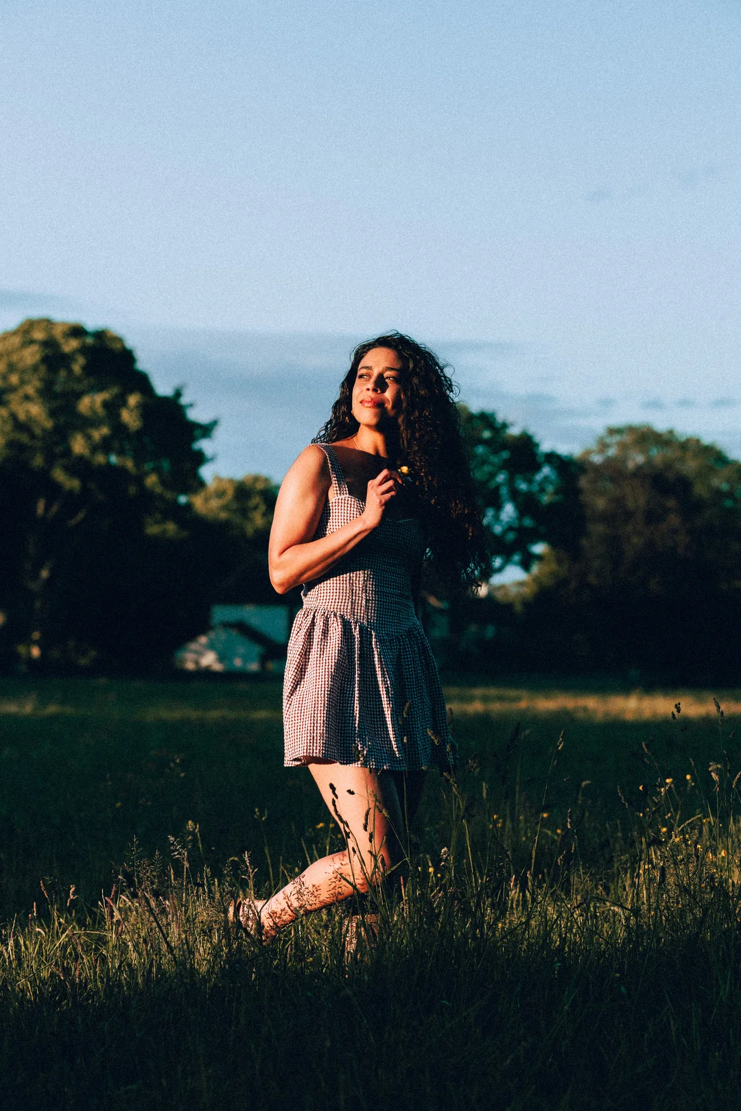 Woman with curly hair in a gingham dress standing in a sunlit grassy field.