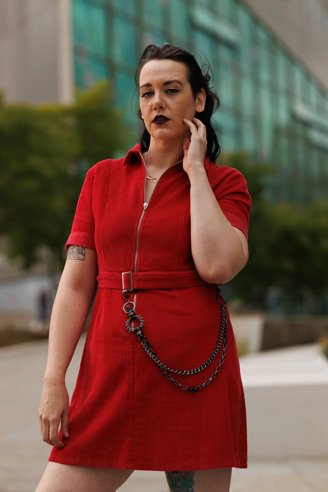 A person with dark hair and makeup wearing a red corduroy mini dress with a chain belt, posing outdoors.