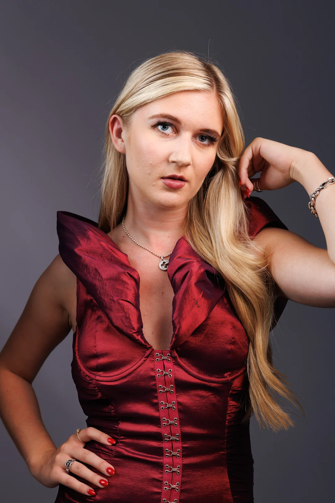A blonde woman wearing a structured red dress posing against a neutral grey studio background.