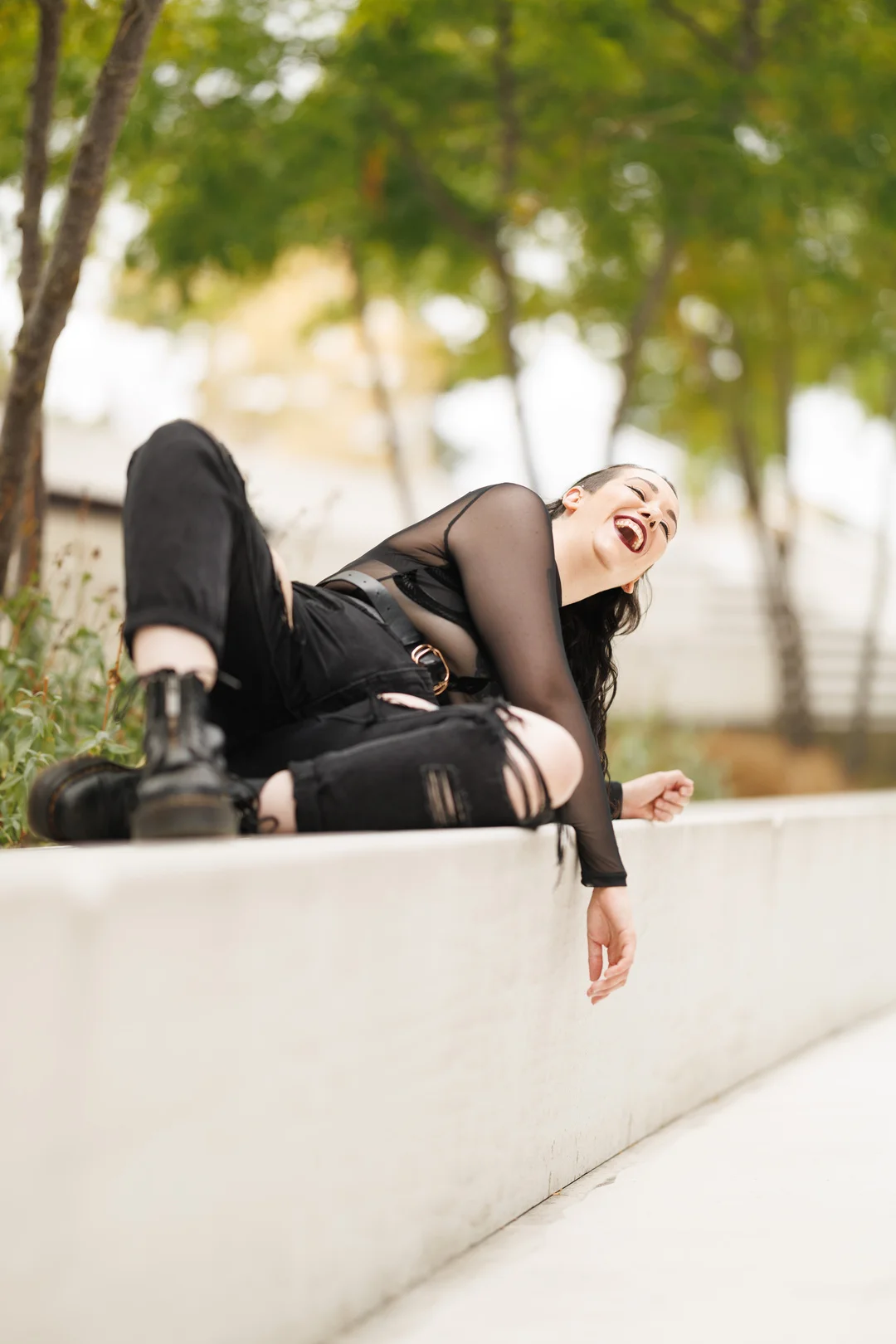 A woman in a black mesh top and ripped black pants laughing while reclining on a white concrete ledge outdoors.