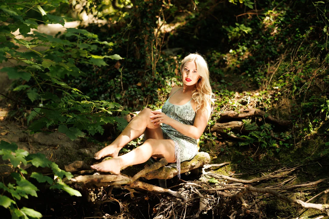 Blonde woman in a light-colored dress posing on a fallen log in a lush green forest.