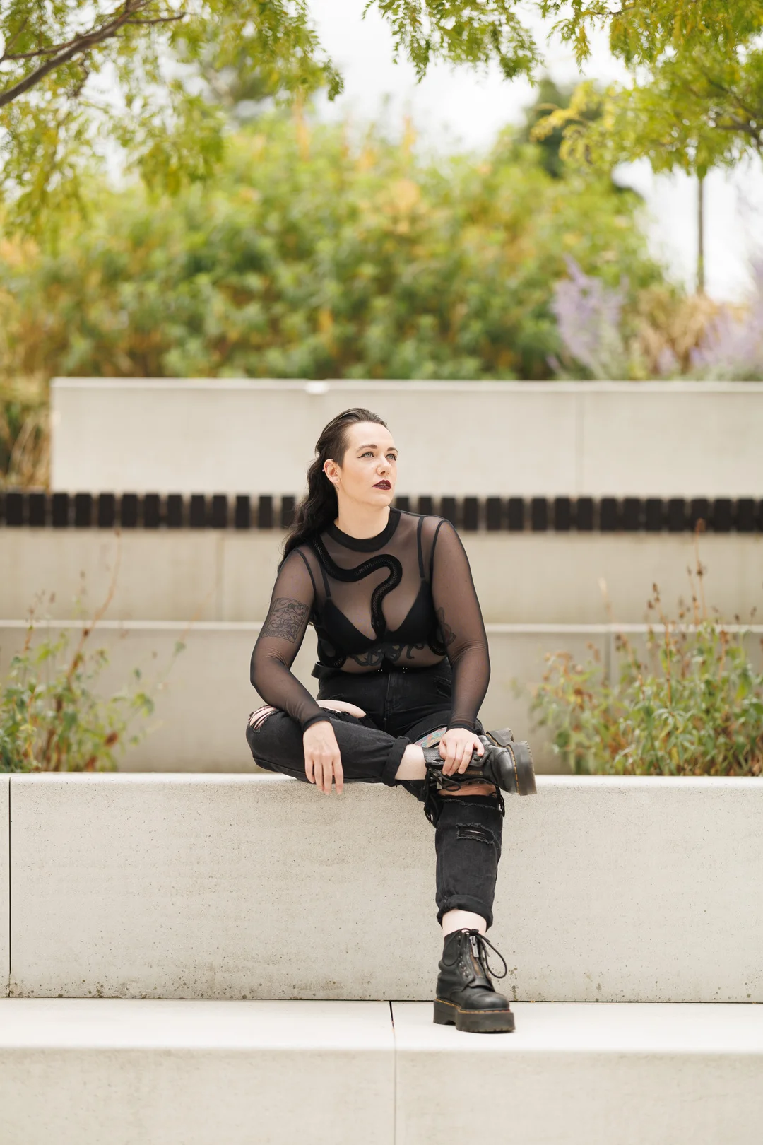 Woman in alternative black fashion sitting on a concrete ledge in an outdoor garden setting.