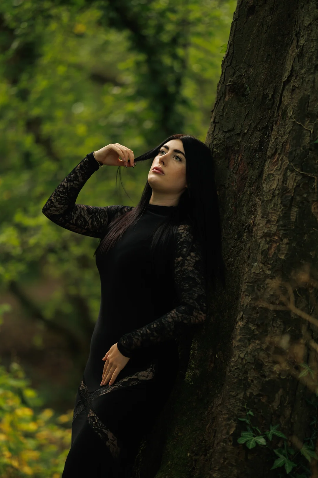 Woman with long black hair in a black lace dress leaning against a tree in a lush green forest.
