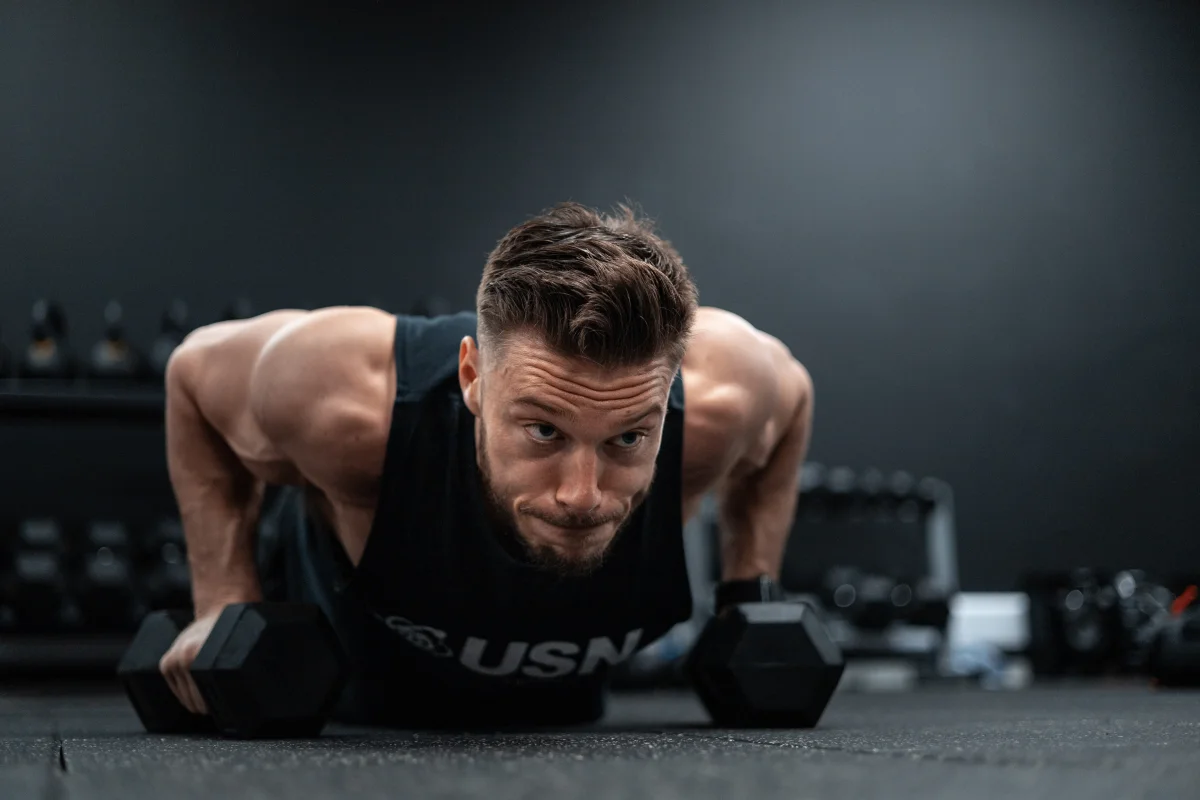 Muscular man in a black tank top performing a push-up on dumbbells in a gym with an intense expression.