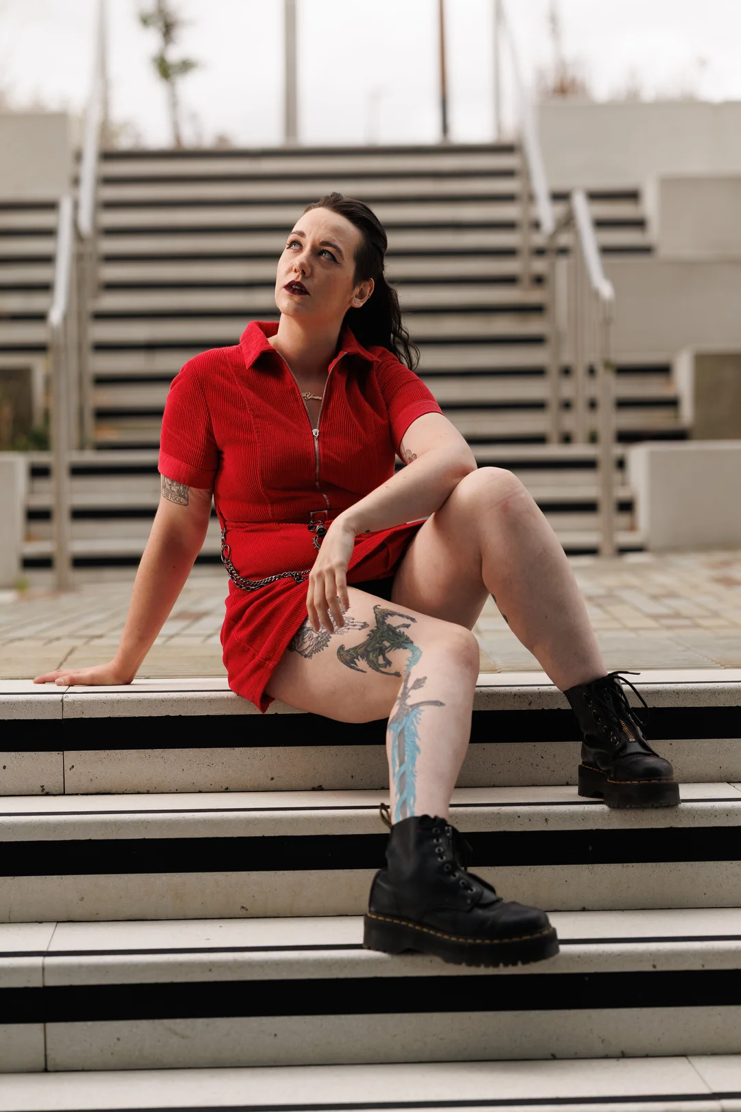 A woman with tattoos wearing a red dress and black boots sitting on outdoor concrete stairs.