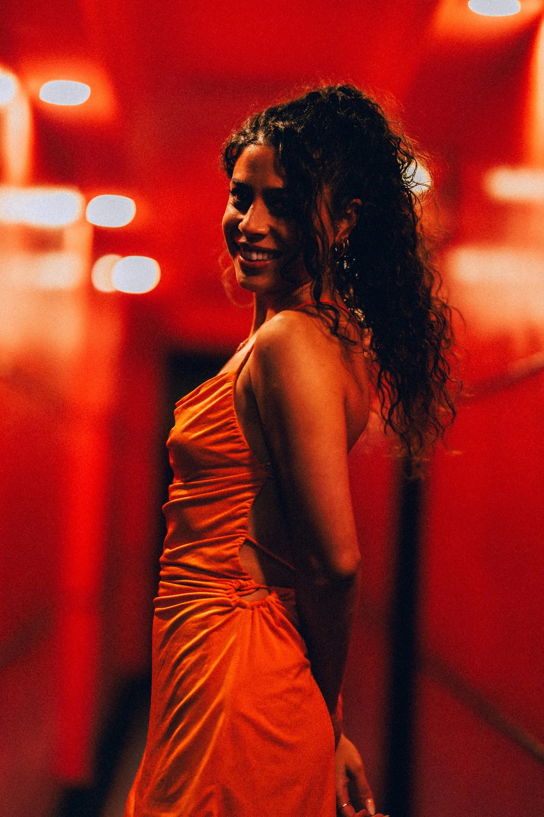Woman with curly hair in an orange dress smiling over her shoulder in a red-lit setting.