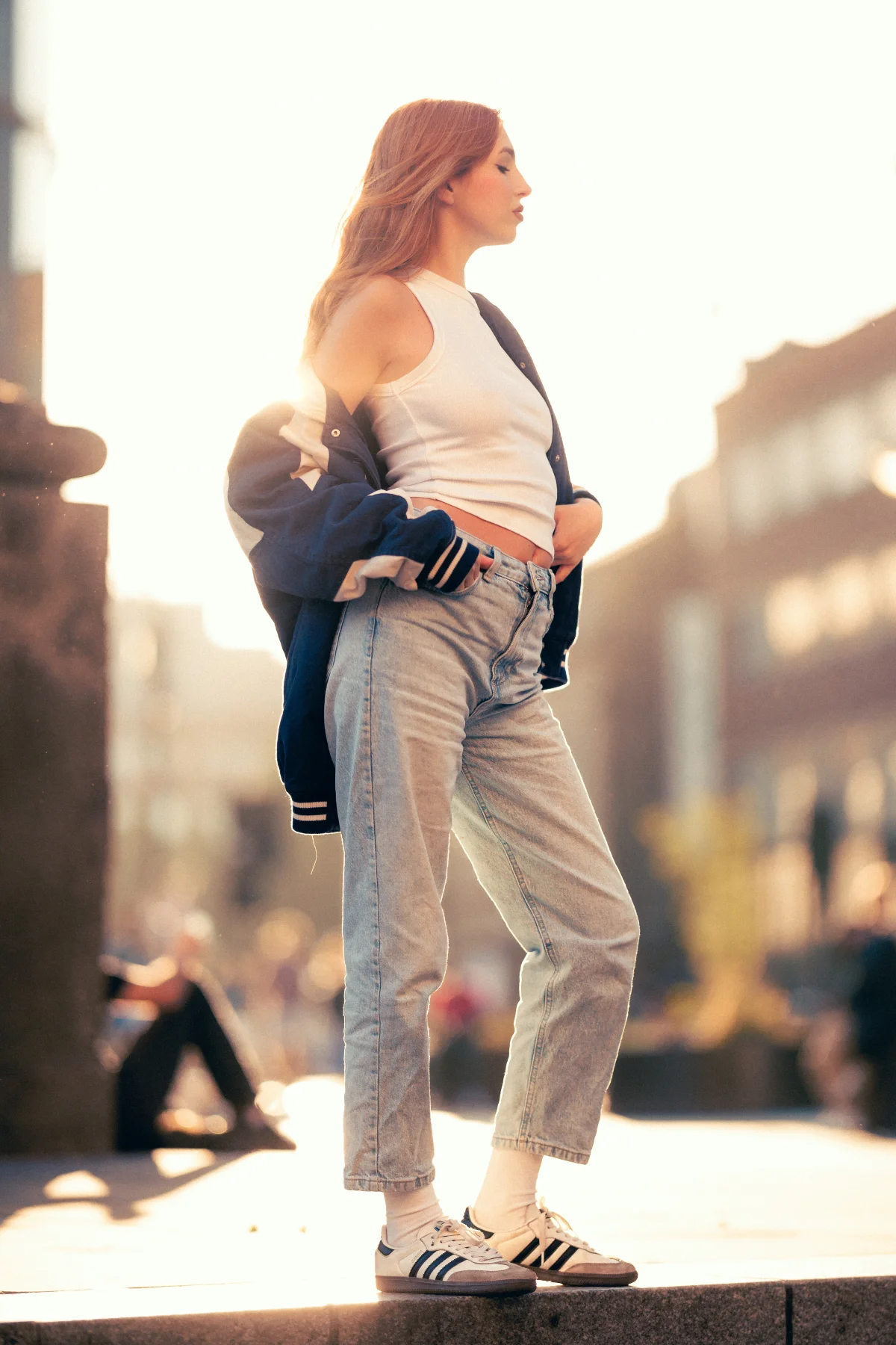 Woman in a white tank top, light-wash jeans, and navy varsity jacket posing in a sunny urban setting.
