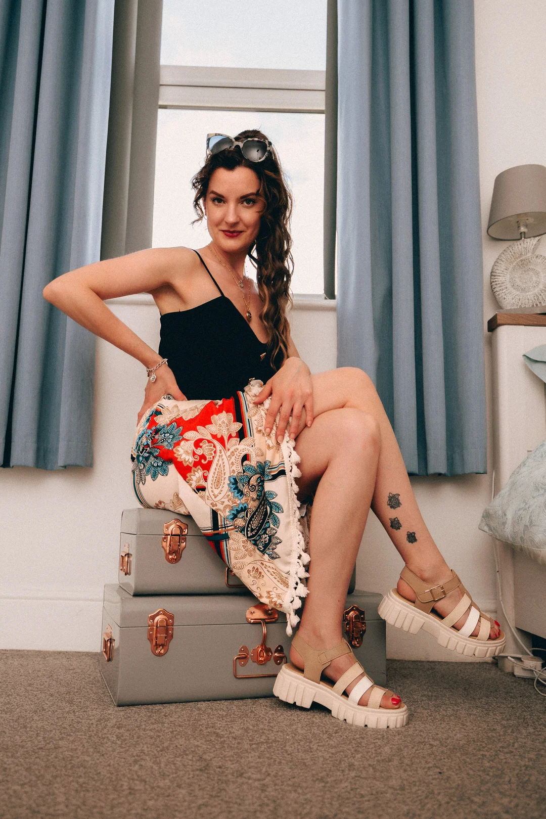 Woman in a black top and colorful patterned skirt posing on grey suitcases by a window.