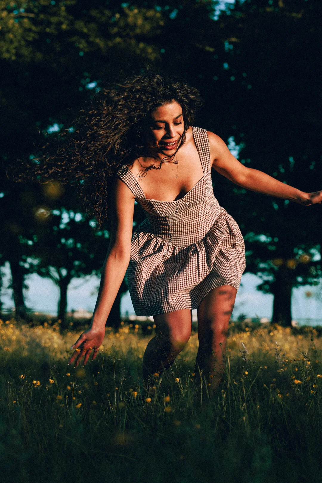 A woman with curly hair in a red and white gingham dress dancing in a field of yellow flowers at sunset.
