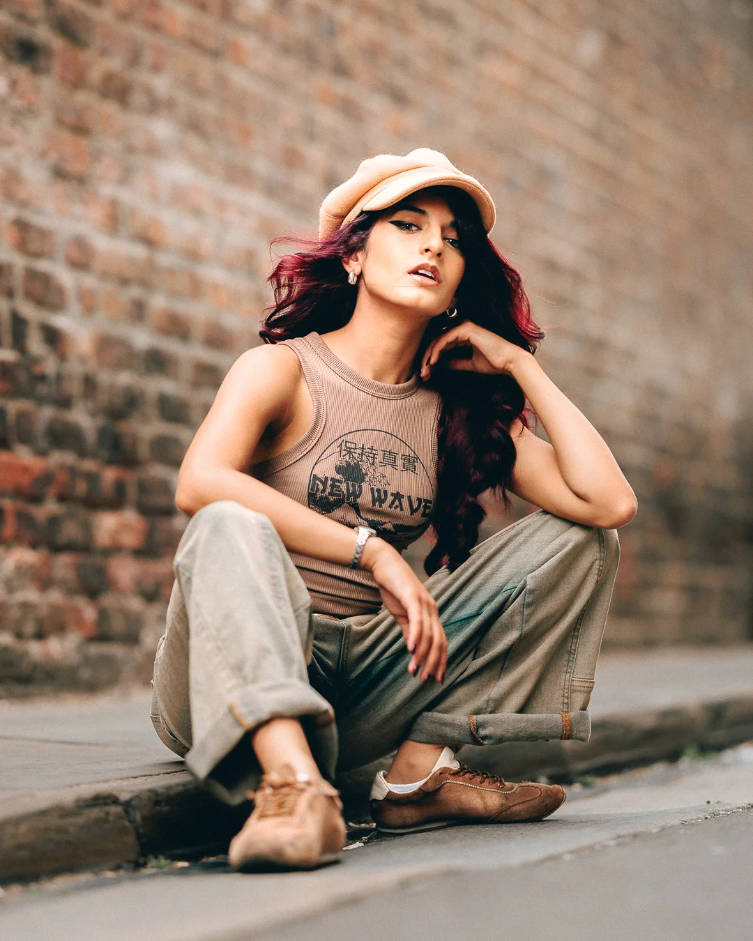 Woman with red hair in a tan cap and tank top crouching against a brick wall in an urban setting.
