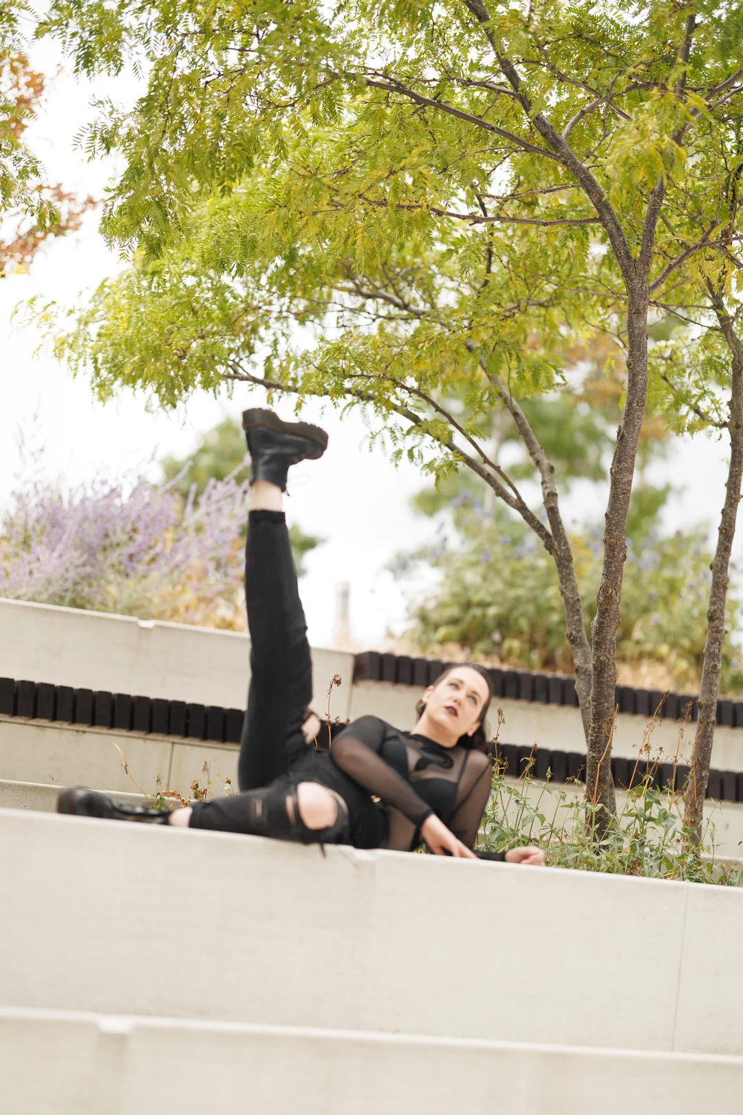 A person in a sheer black top and ripped jeans posing creatively on a concrete ledge under a green tree.