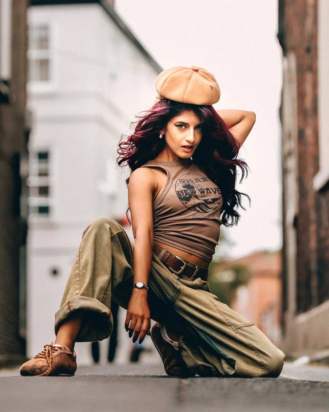 Woman with burgundy hair in a tan beret and olive cargo pants posing in an urban alleyway.