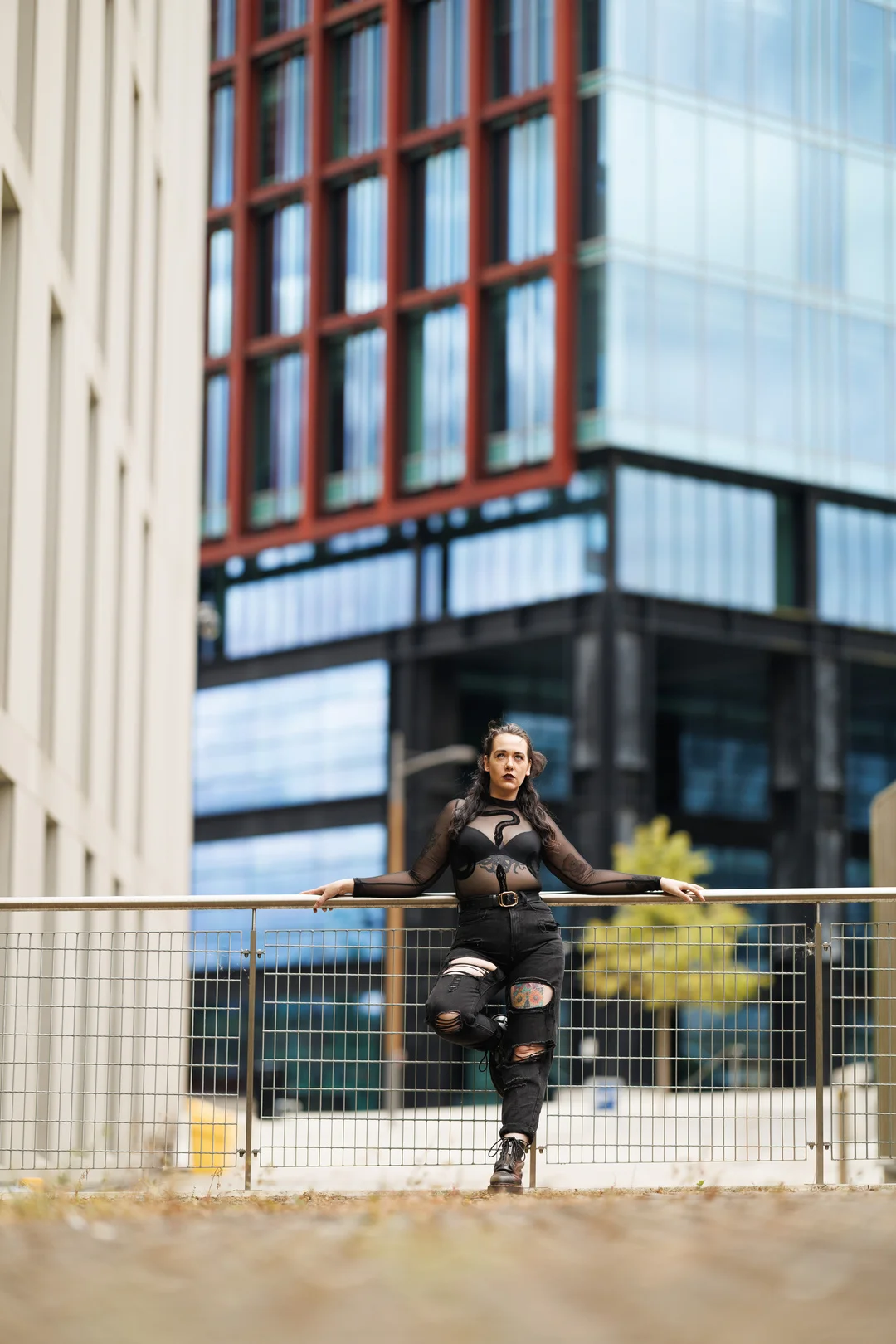 Woman in a black sheer top and ripped black jeans posing by a railing with modern buildings behind her.