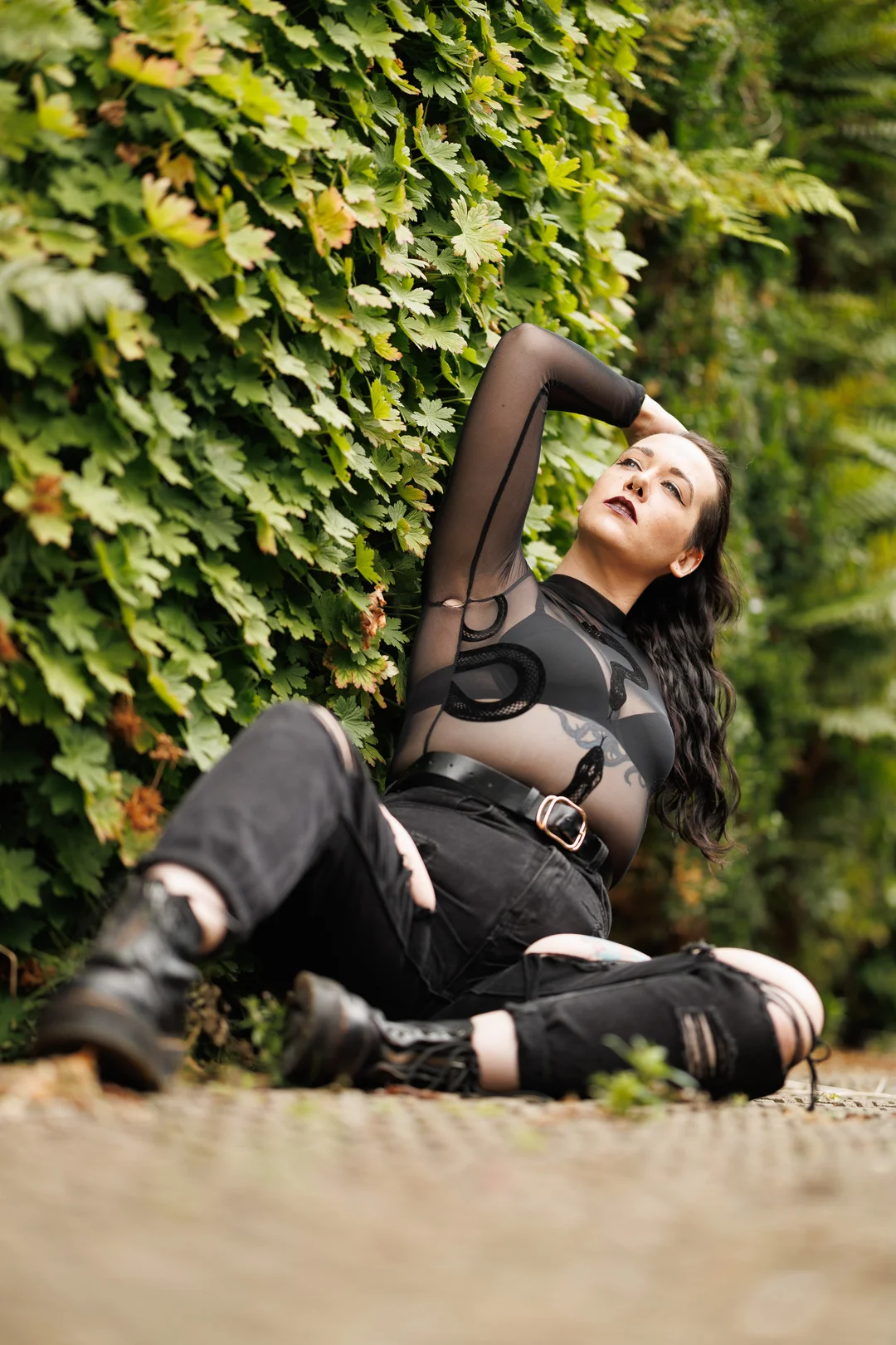 Woman in a sheer black top and ripped jeans posing against a lush green hedge.