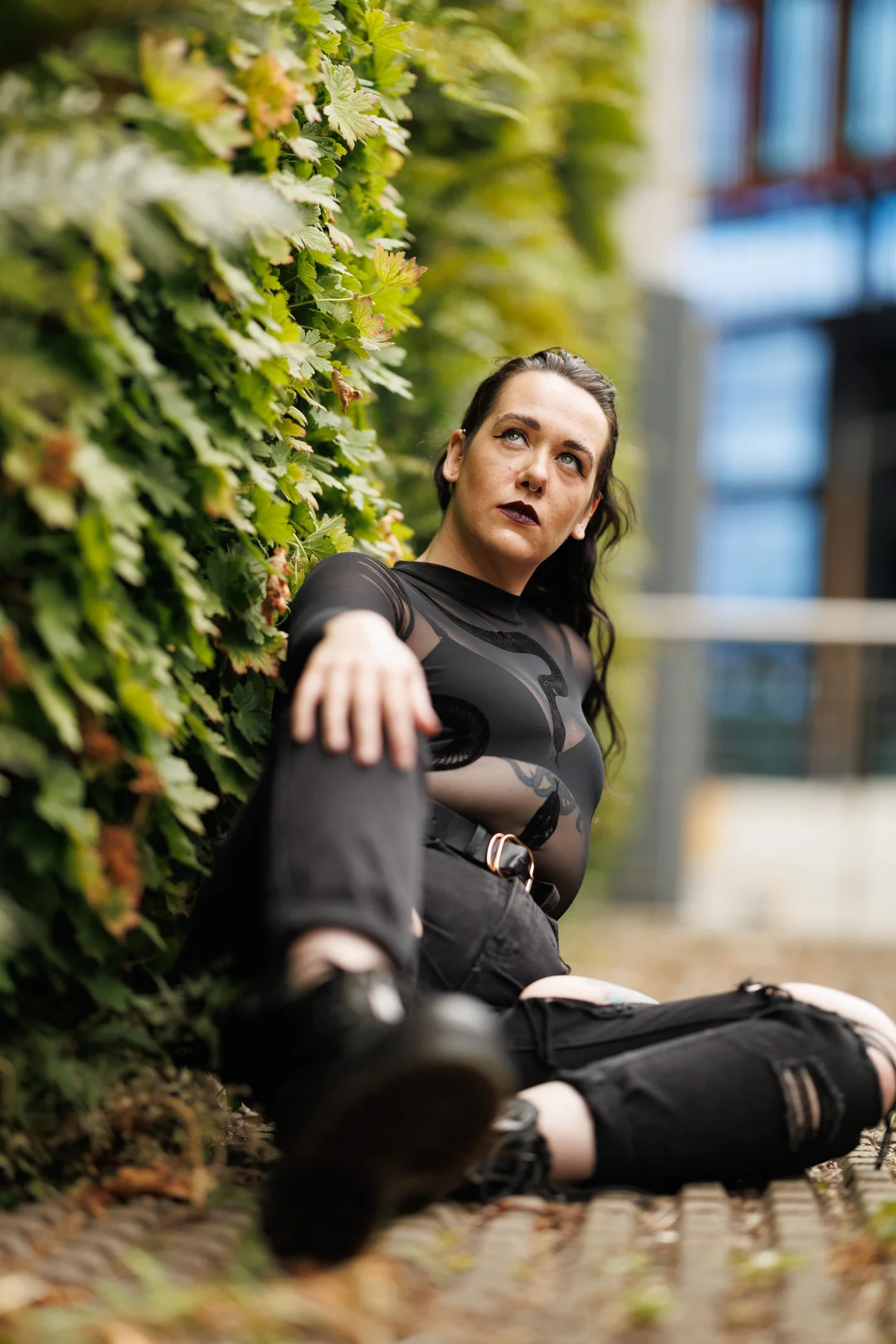 Person with dark hair and makeup in a sheer black top and ripped jeans sitting against a green hedge.