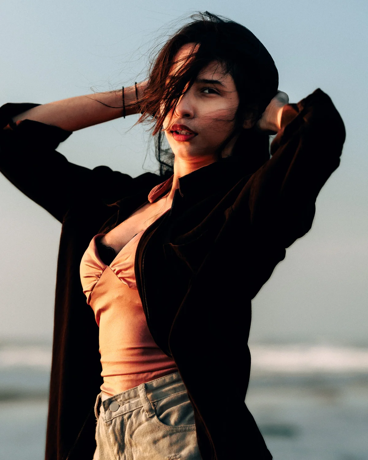 A woman with dark hair posing with hands behind her head, wearing a black jacket and peach top in soft outdoor light.