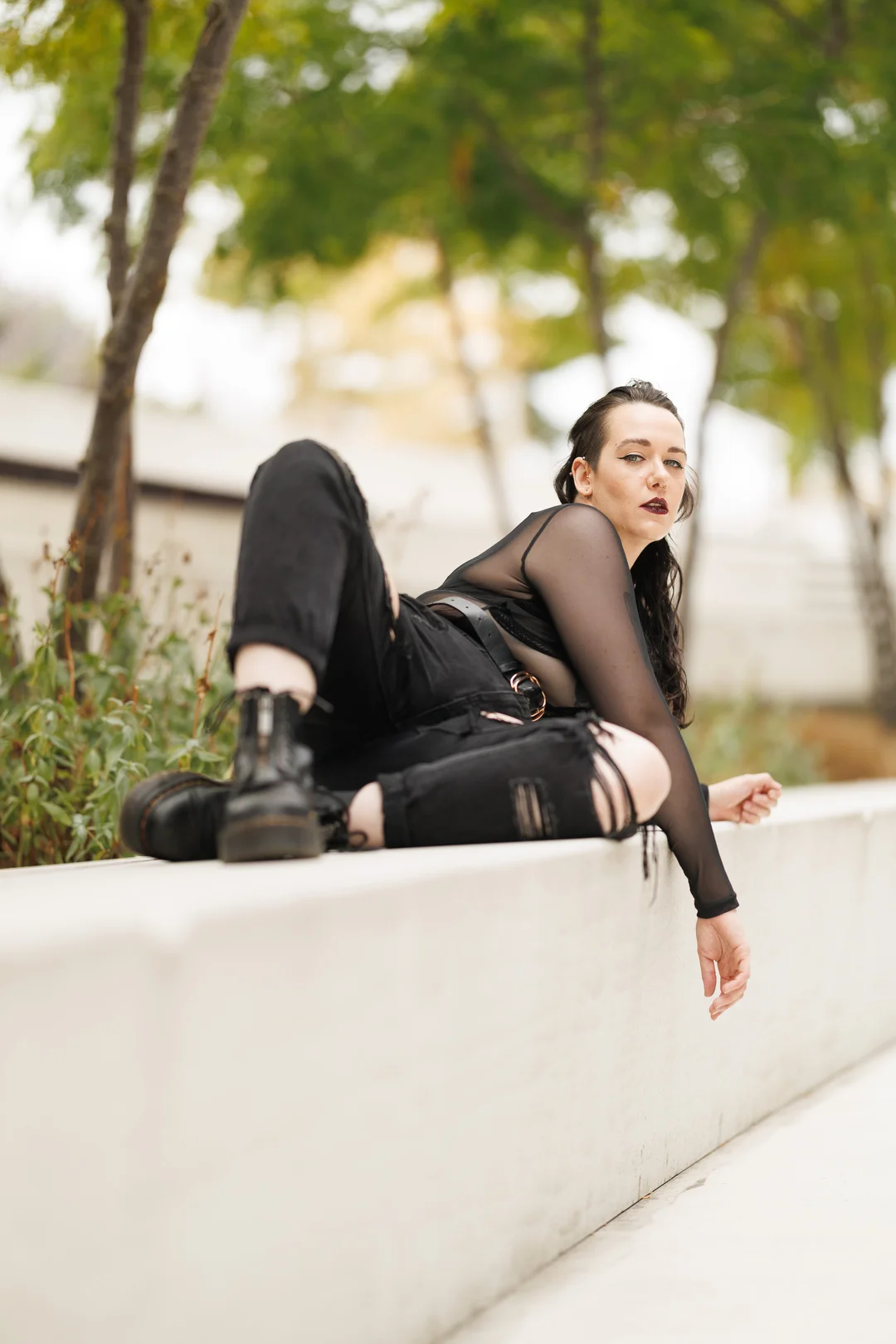Person in alternative black attire posing on a white concrete ledge in an outdoor setting.