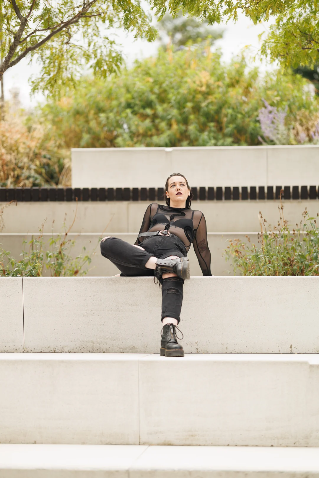 A person with dark makeup and an edgy black outfit posing on concrete steps in a garden.