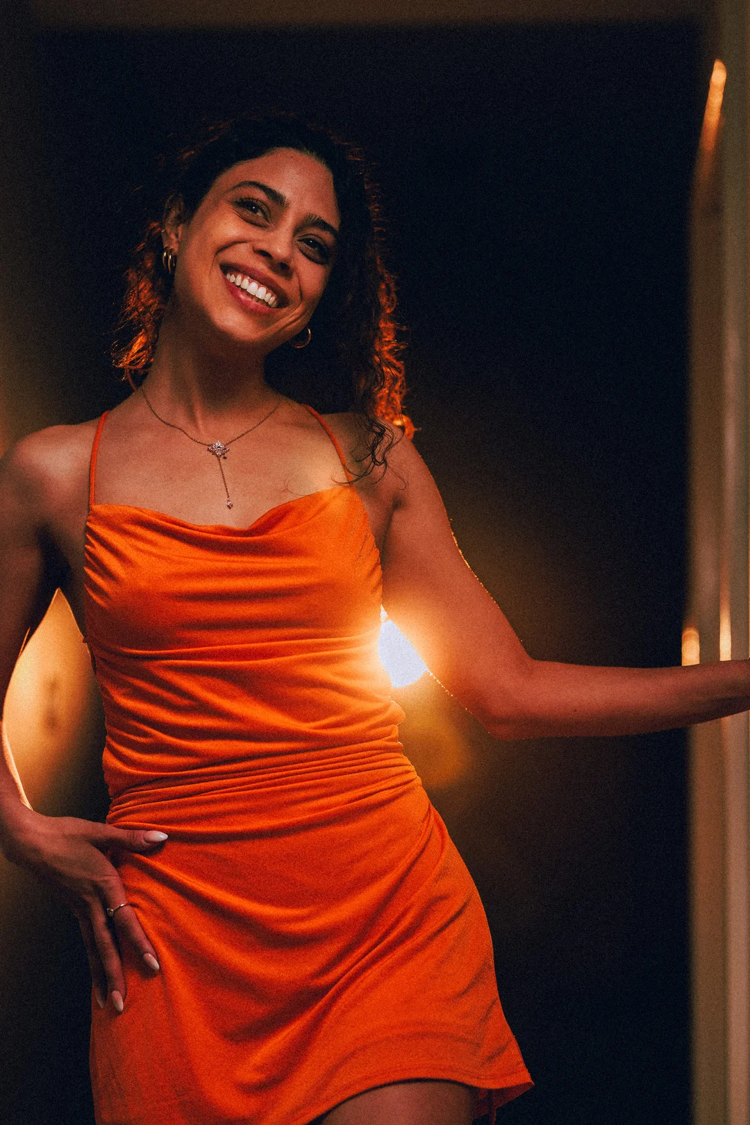 Smiling woman with curly hair in an orange satin dress, illuminated by warm backlighting.