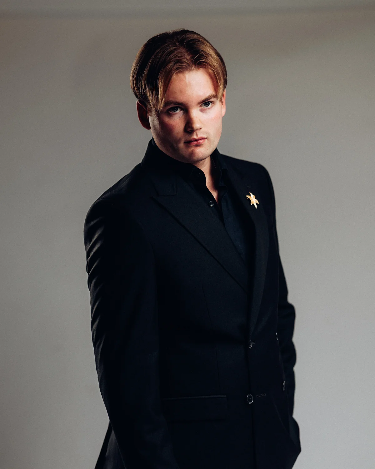 A young man with reddish-blonde hair in a black suit and shirt, posing against a neutral gray studio background.