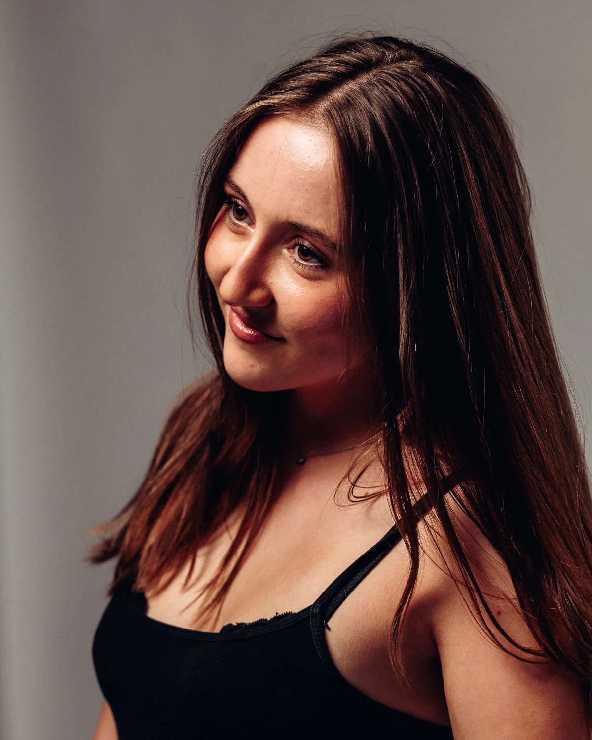 Close-up portrait of a young woman with long brown hair and a slight smile against a neutral background.