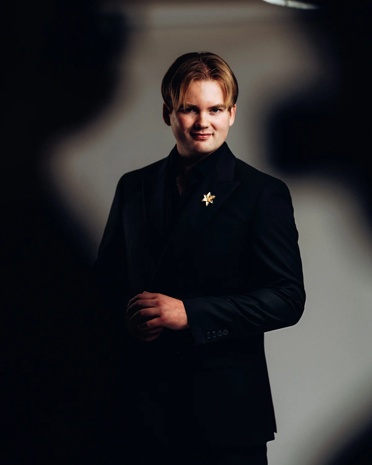A man in a black suit with a gold flower pin, posing against a dark, moody background.