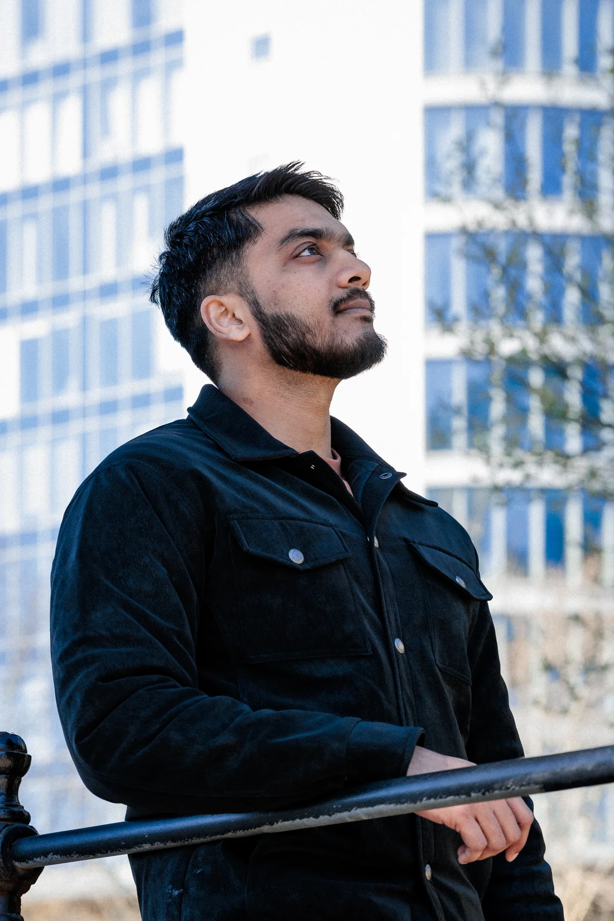 Side profile portrait of a bearded man in a black jacket against a blurred urban city background.