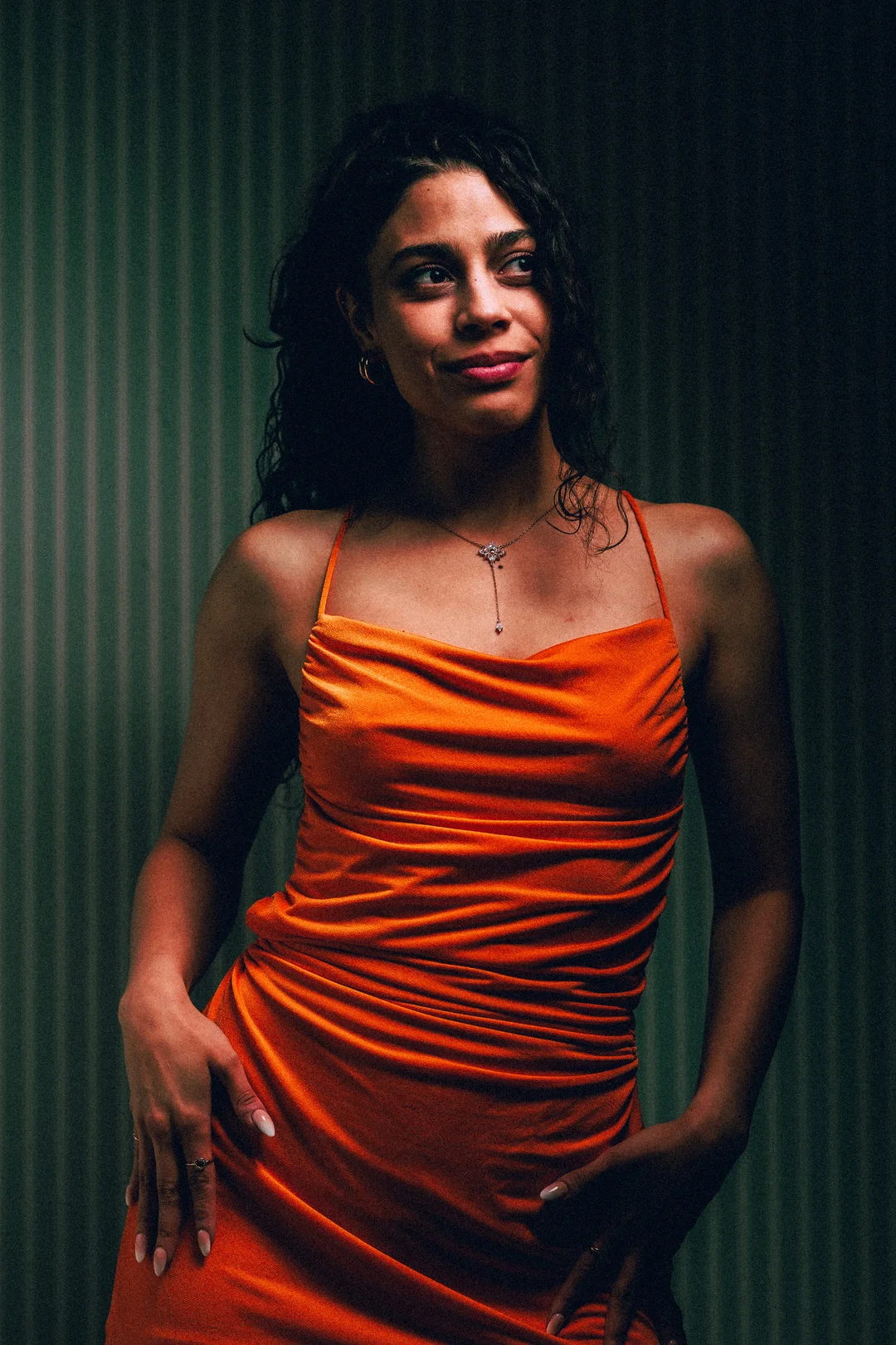 A woman with curly dark hair in an orange satin dress posing against a dark green striped background.