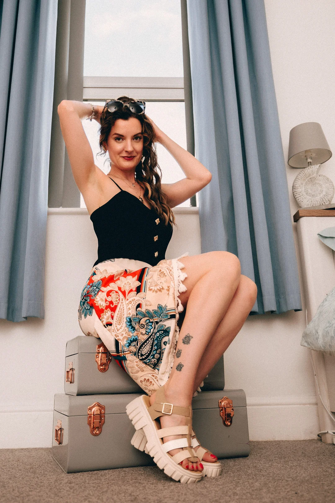 A woman in a black top and paisley skirt posing while sitting on grey suitcases in front of a window.