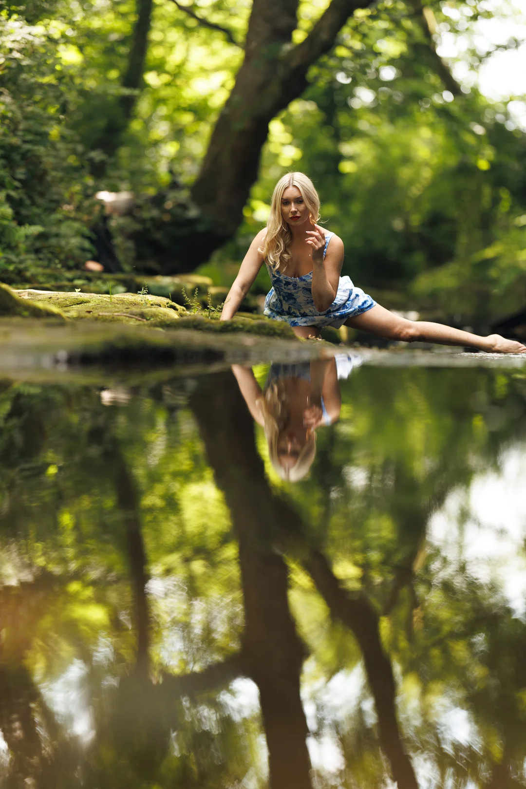 Blonde woman in a blue floral dress posing by a forest stream with a clear reflection in the water.