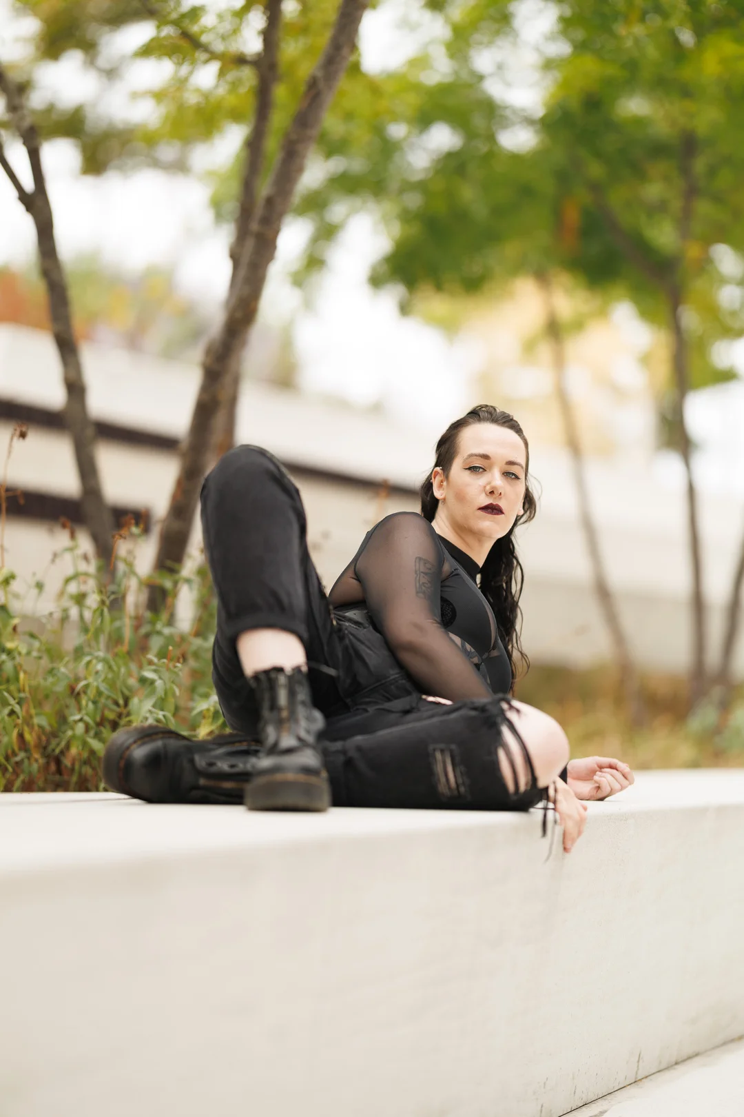 A woman in black alternative fashion sitting on a white concrete ledge in an outdoor setting.