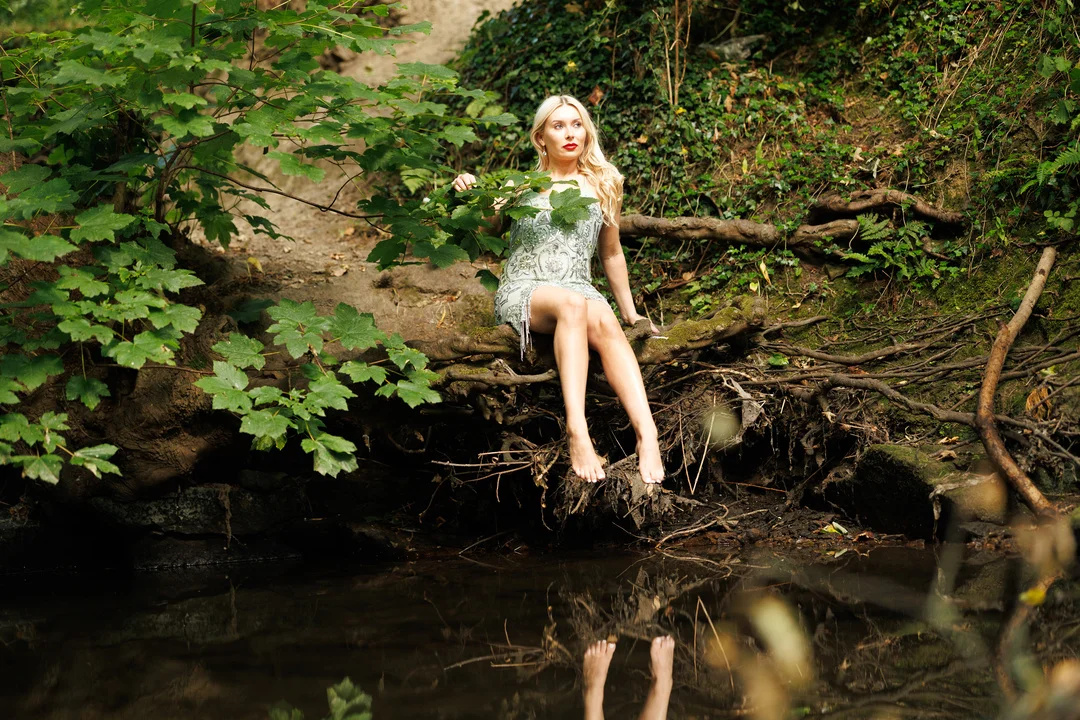 Blonde woman in a white lace dress sitting on a riverbank in a lush green forest.