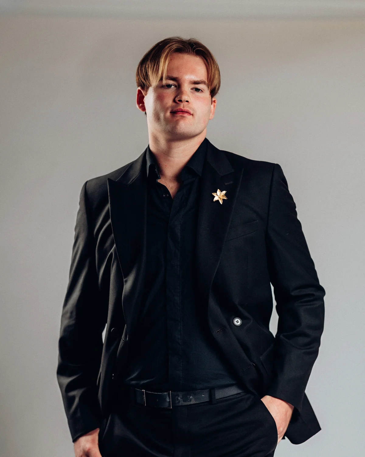 A man with blond hair in a black suit and shirt posing against a neutral grey studio background.