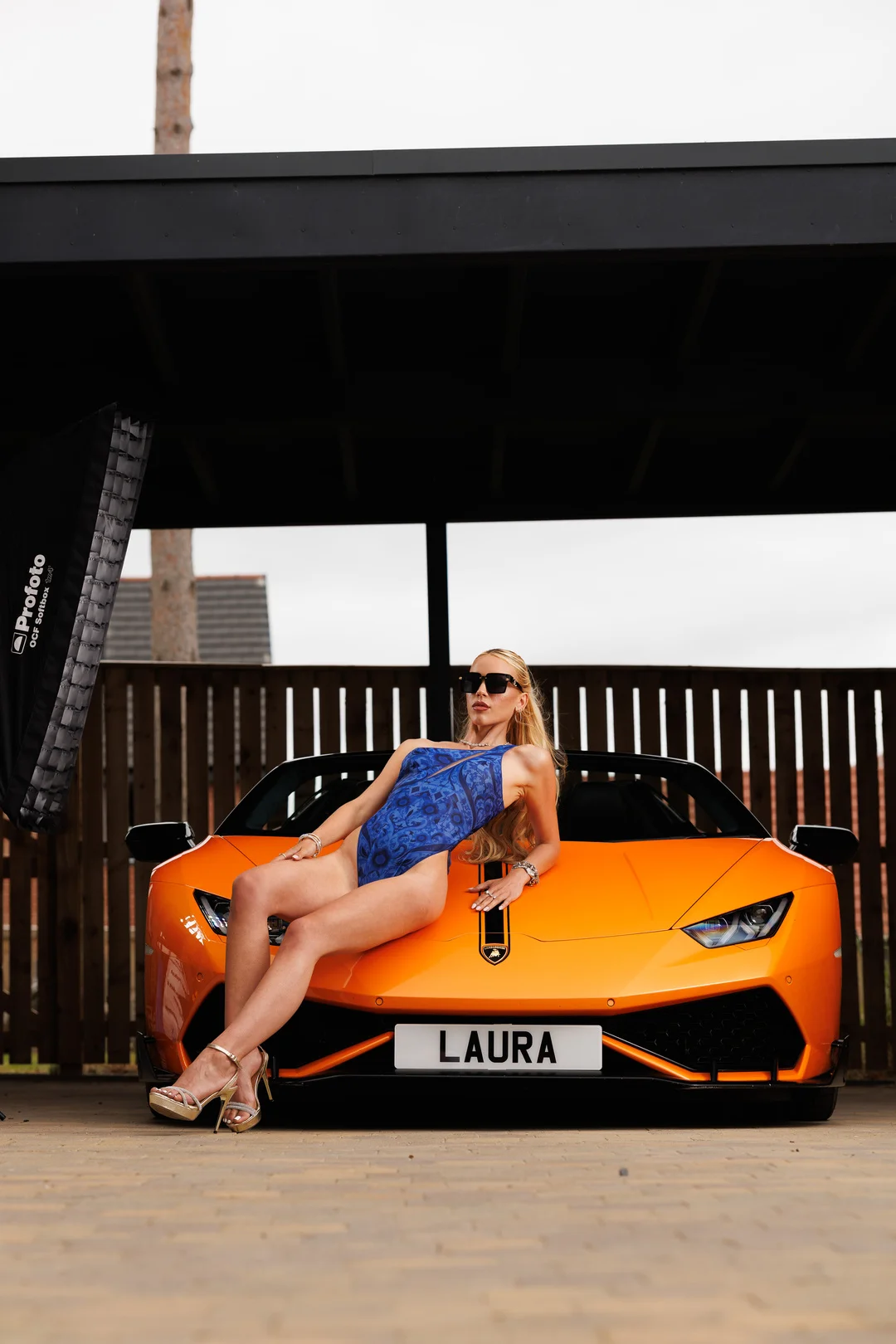 Blonde woman in a blue swimsuit and sunglasses posing on the hood of an orange Lamborghini.