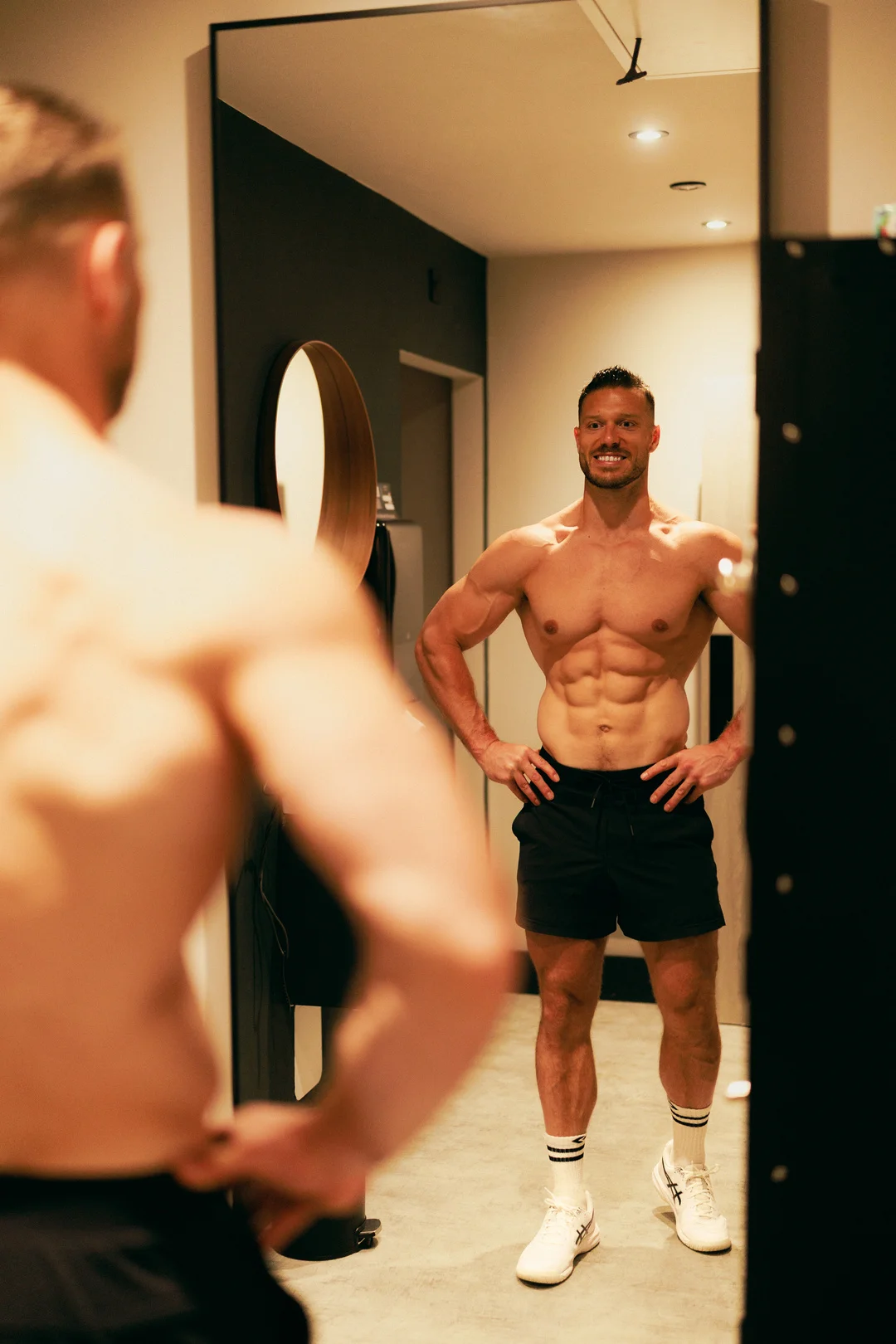 Muscular man smiling at his reflection in a mirror, wearing black athletic shorts and white sneakers.